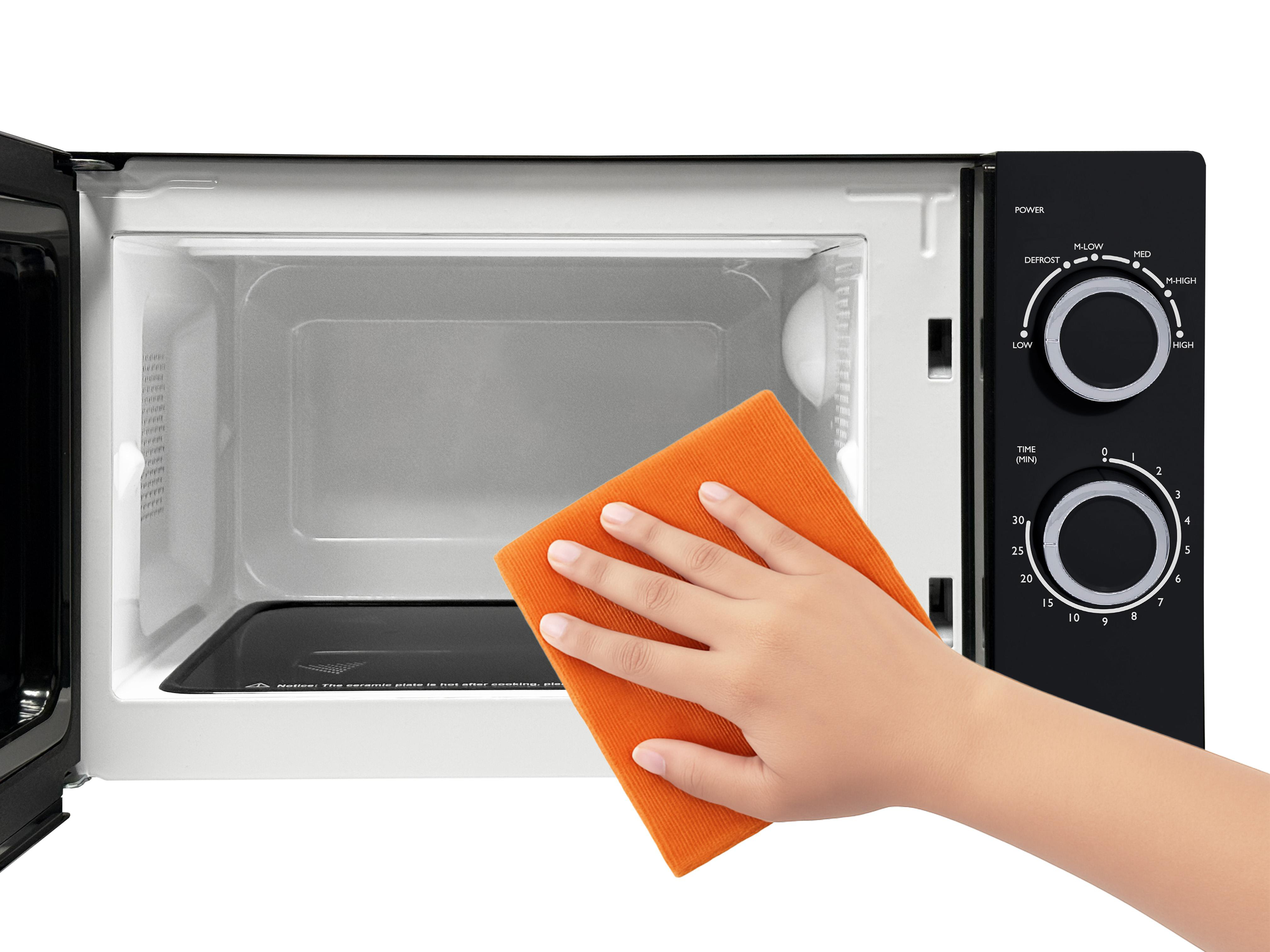 Eine Hand reinigt eine Mikrowelle mit einem orangefarbenen Tuch. Die Mikrowelle ist geöffnet. Bedienfeld ist rechts.