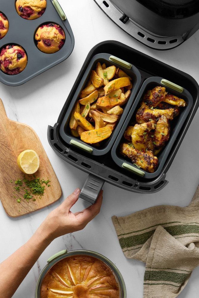 An air fryer with roasted potatoes and chicken wings. A wooden board with lemon and parsley. Muffins and cake are also present.
