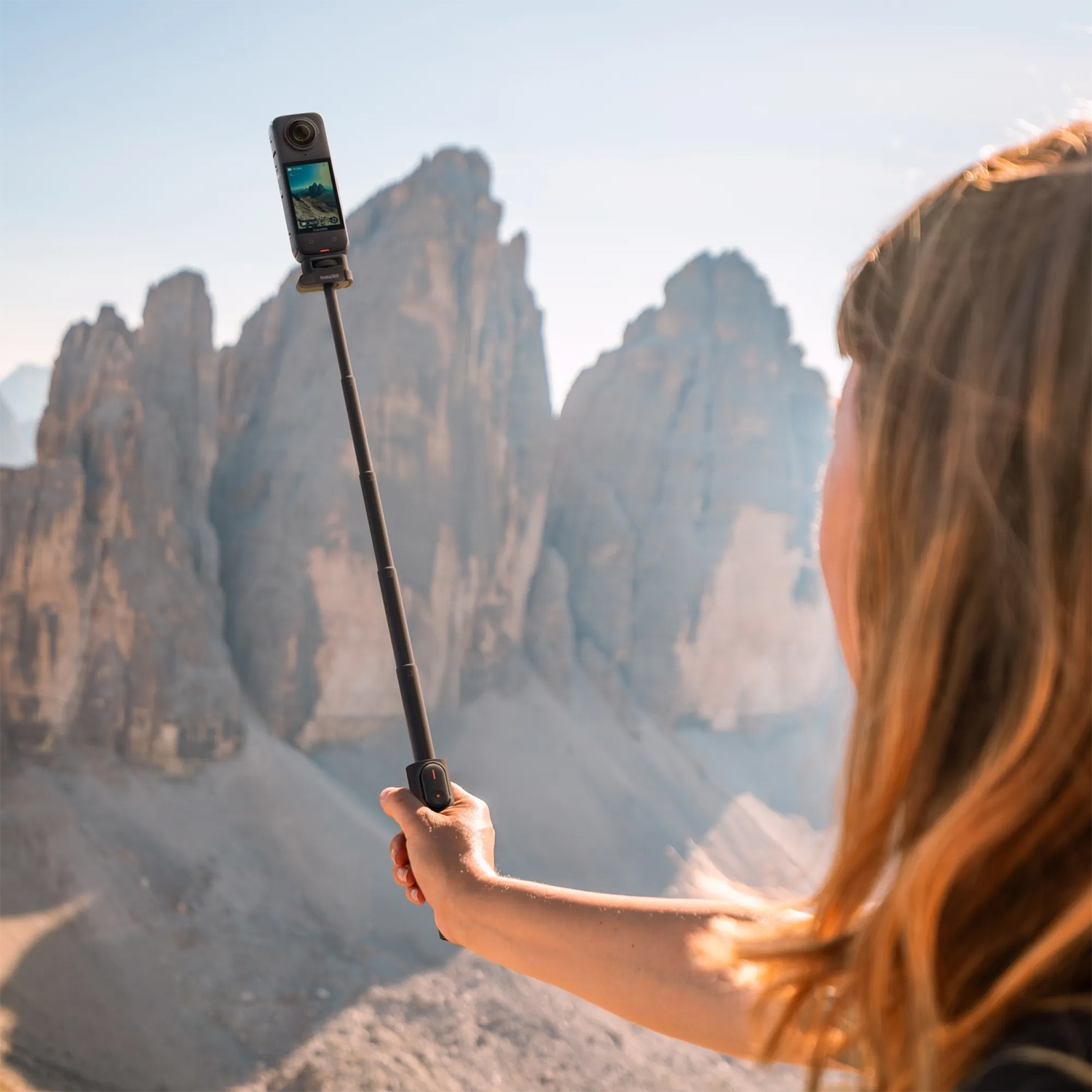 Una persona tiene una fotocamera su un bastone per selfie, filmando montagne. La fotocamera mostra la vista. I capelli sono dorati.