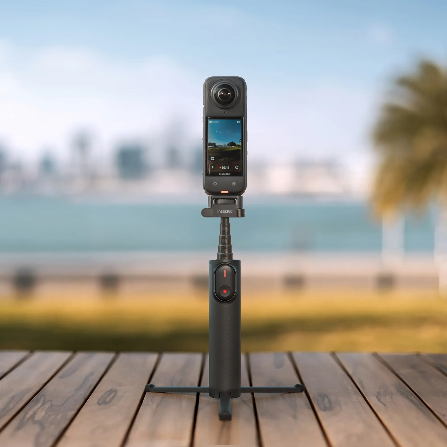 Una fotocamera nera su un treppiede, che mostra uno schermo con vista paesaggistica. Lo sfondo è un paesaggio urbano sfocato vicino al mare.