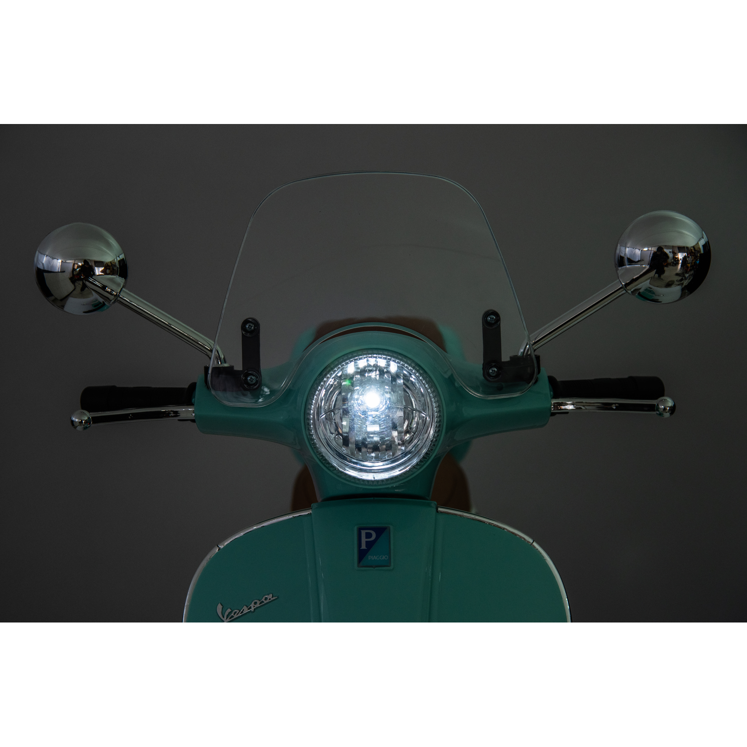 Front view of a teal Vespa scooter. It has a windshield, chrome mirrors, and a bright headlight. The background is a gradient gray.