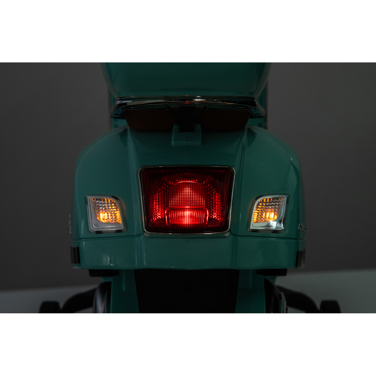 Rear view of a teal scooter, with a bright red tail light and amber turn signals. The background is a dark gray.