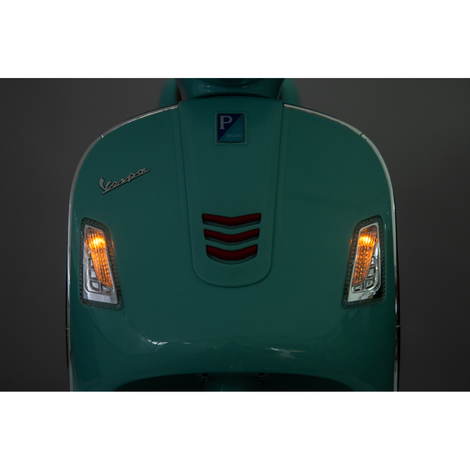 Close-up of a teal Vespa scooter. The Vespa logo is visible. Turn signals are illuminated orange. The background is dark gray.