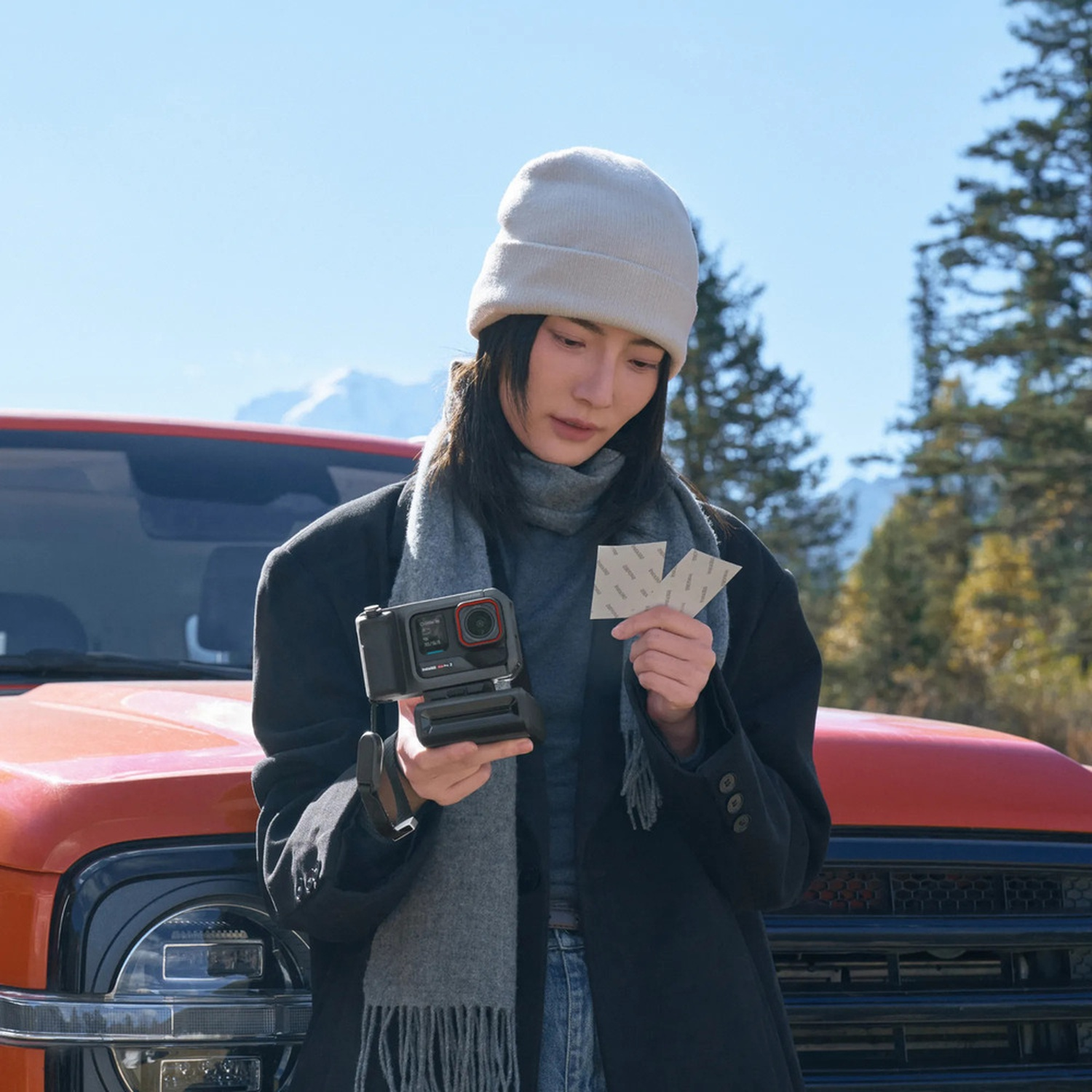 Una persona tiene in mano una fotocamera e delle carte vicino a un camion arancione, indossa un berretto, una sciarpa e un cappotto, con alberi e montagne sullo sfondo.
