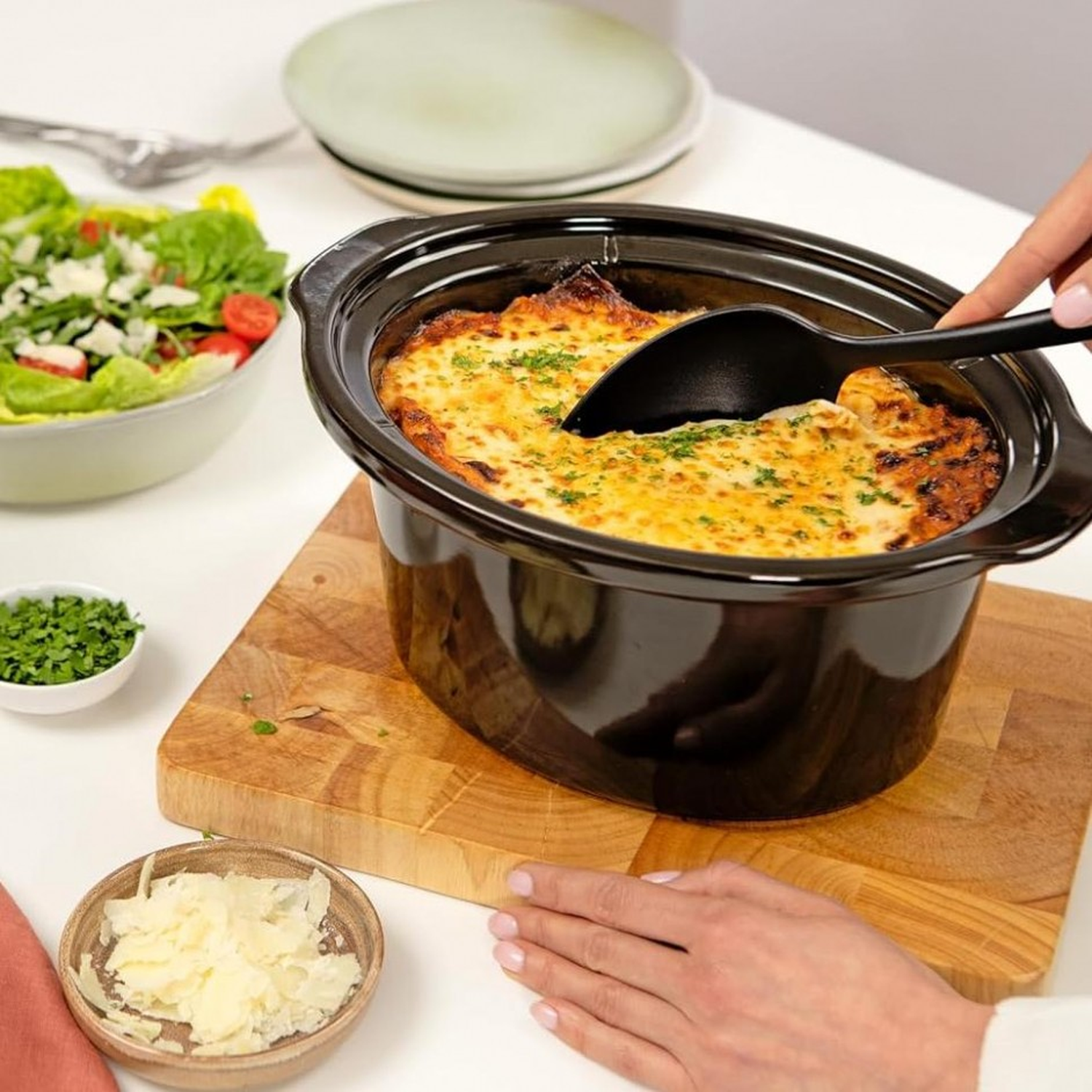 A black pot with baked dish. A salad and cheese are on the table. A hand is scooping food with a black spoon.