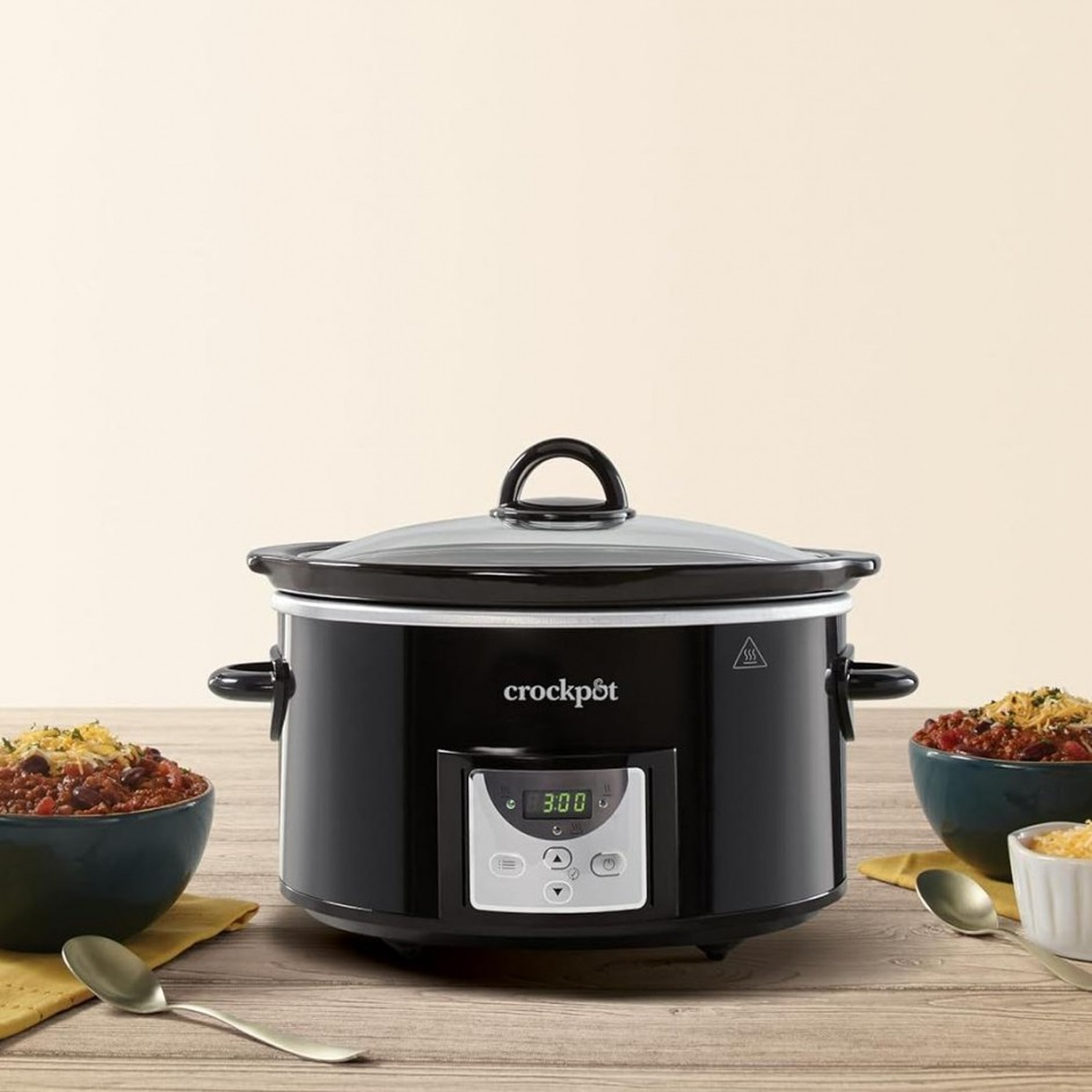 A black Crock-Pot with a glass lid sits on a wooden table. Bowls of chili and spoons are placed around it.