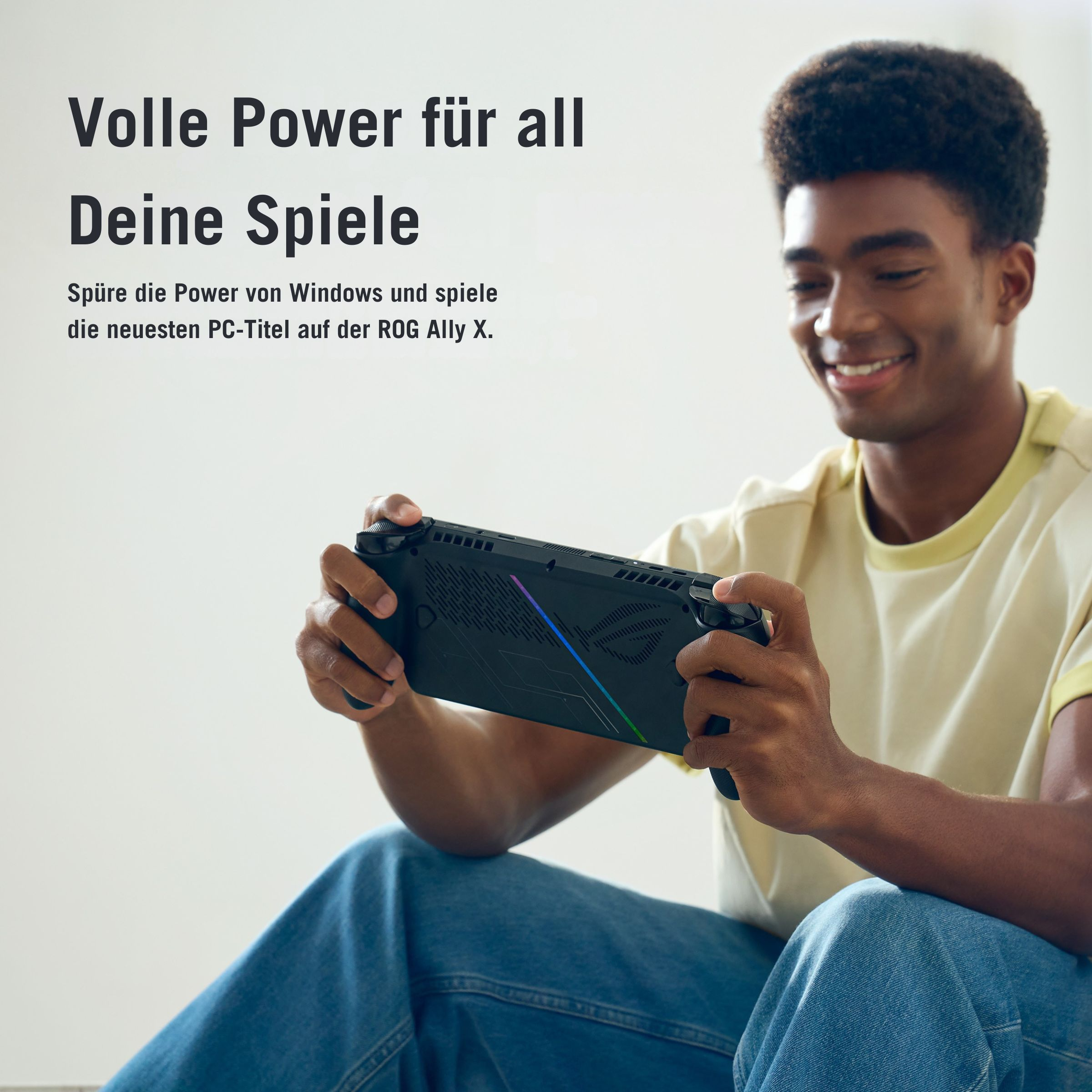 Ein Mann hält lächelnd ein schwarzes Handheld-Gaming-Gerät. Das Gerät hat bunte Lichter. Er trägt ein gelbes Hemd und Jeans.