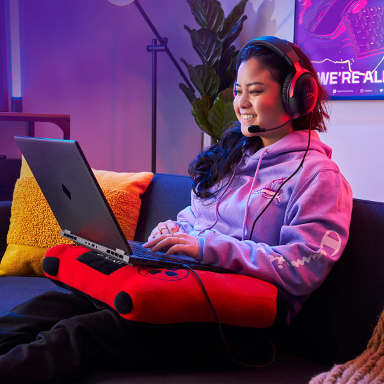 Mujer con auriculares usando laptop en sofá. Cojín rojo soporta la laptop. Vistiendo sudadera morada, sonriendo a la pantalla.