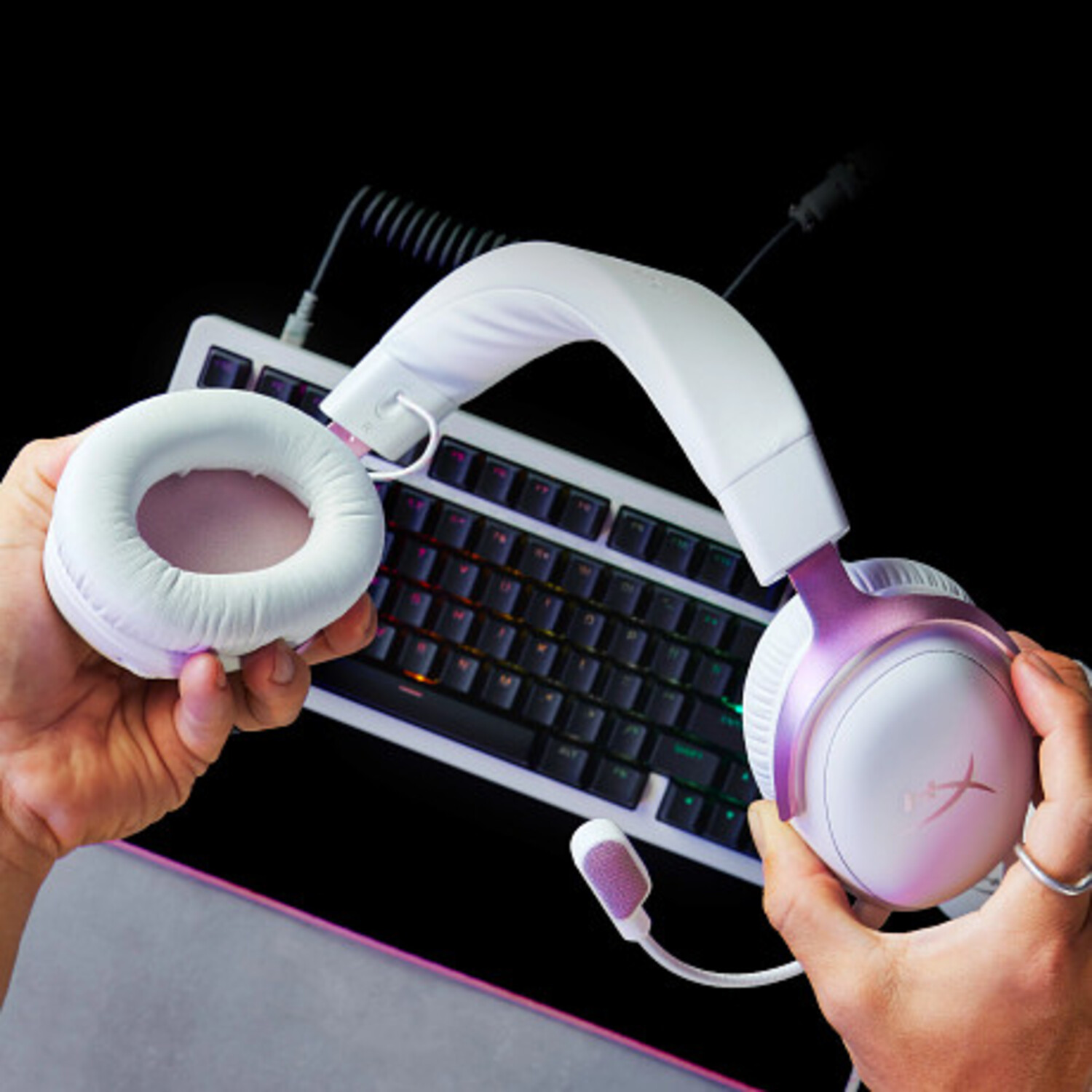 Manos sosteniendo auriculares para juegos blancos y morados con un teclado en el fondo. Fondo negro.