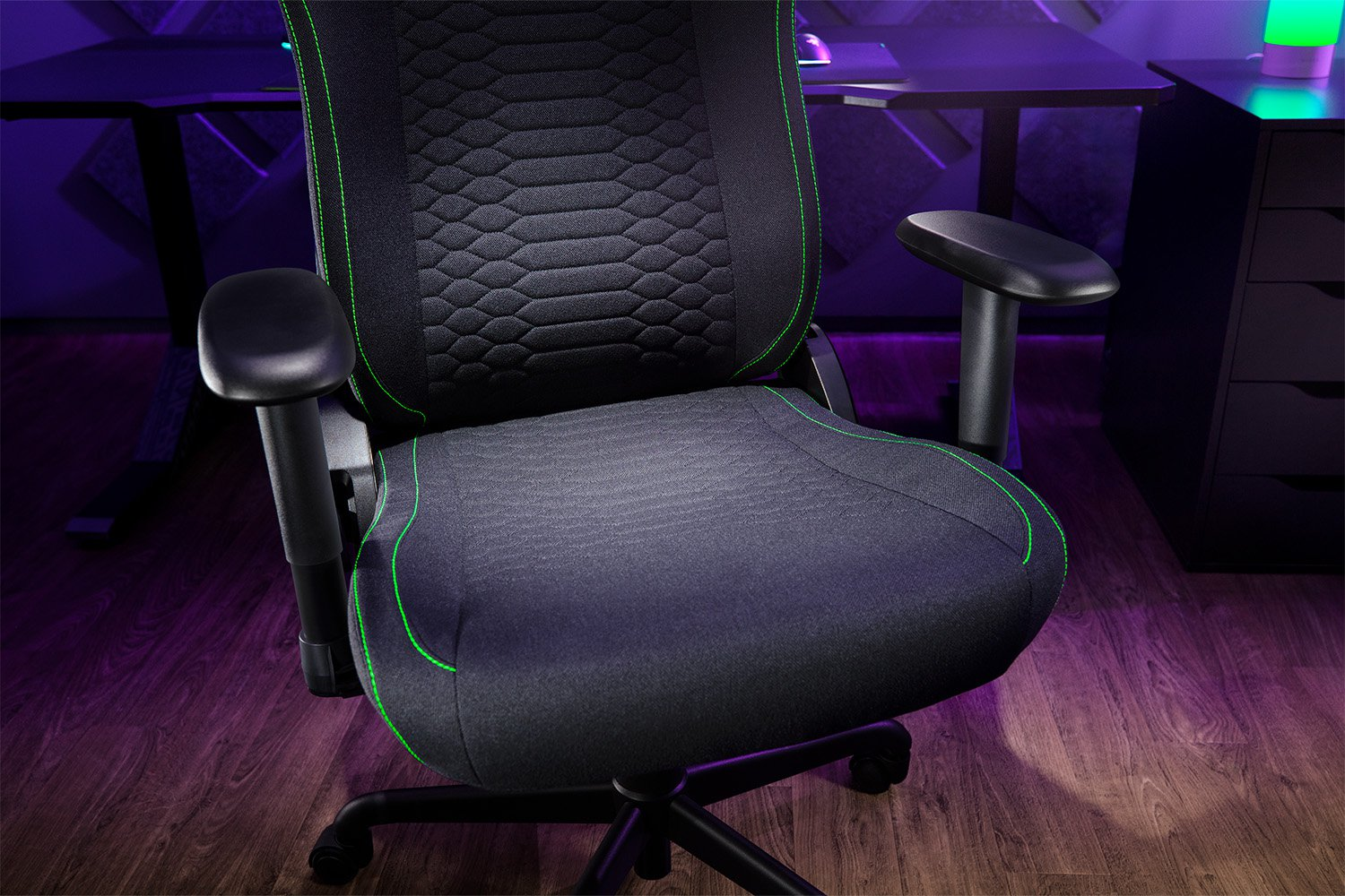 Fekete gamer szék zöld varrással. A háttérben egy asztal és egy színes lámpa látható.