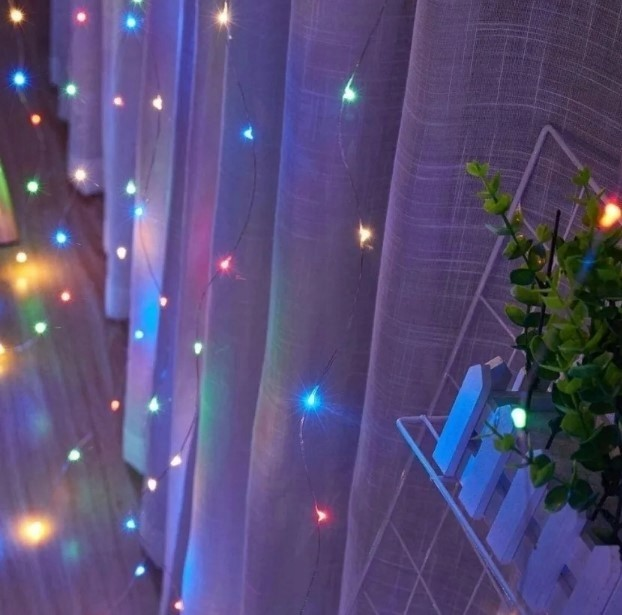 Światła sznurkowe z kolorowymi światłami na tle białych zasłon. Mała roślina w prawym rogu.