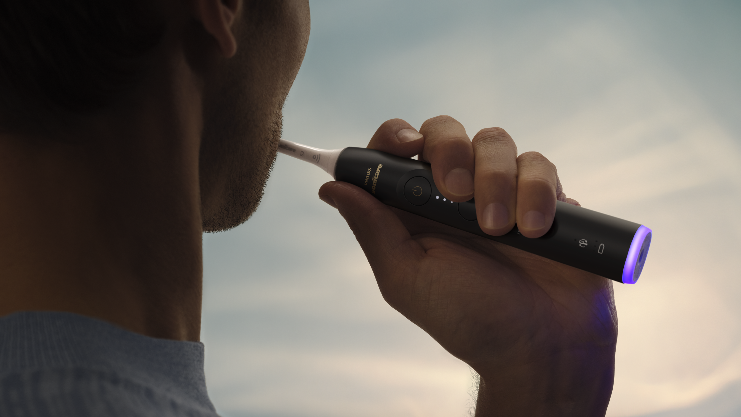 Une personne utilise une brosse à dents Philips Sonicare noire. Le fond montre un ciel bleu et la barbe de la personne.