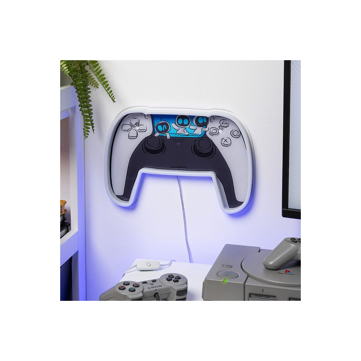 Una luz con forma de mando montada en la pared. Un mando y una consola Playstation están sobre un escritorio blanco. Una planta está en un estante.