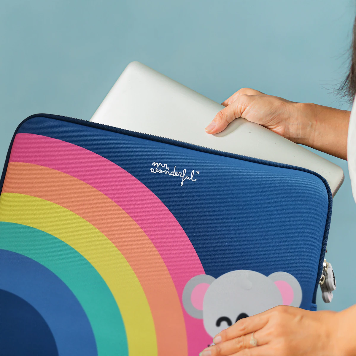 Una persona sostiene una laptop. La laptop se está colocando en una funda azul con diseño de arcoíris e ilustración de koala.