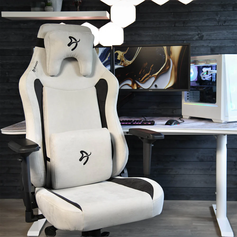 Egy fehér-fekete gamer szék a képen. Egy asztal számítógéppel és monitorral a háttérben.