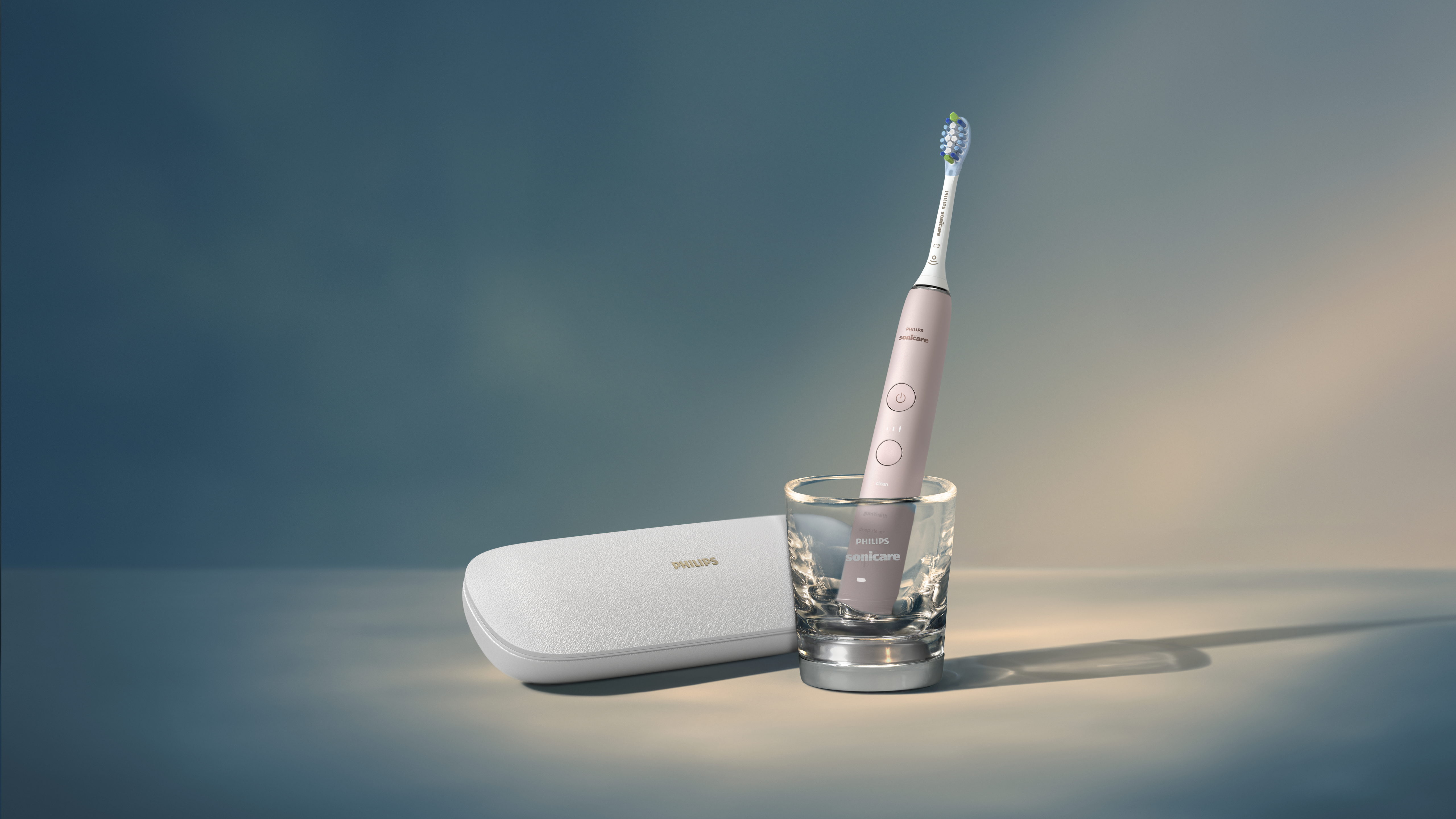 Brosse à dents électrique rose dans un verre, avec un étui blanc. Le fond est turquoise et beige.