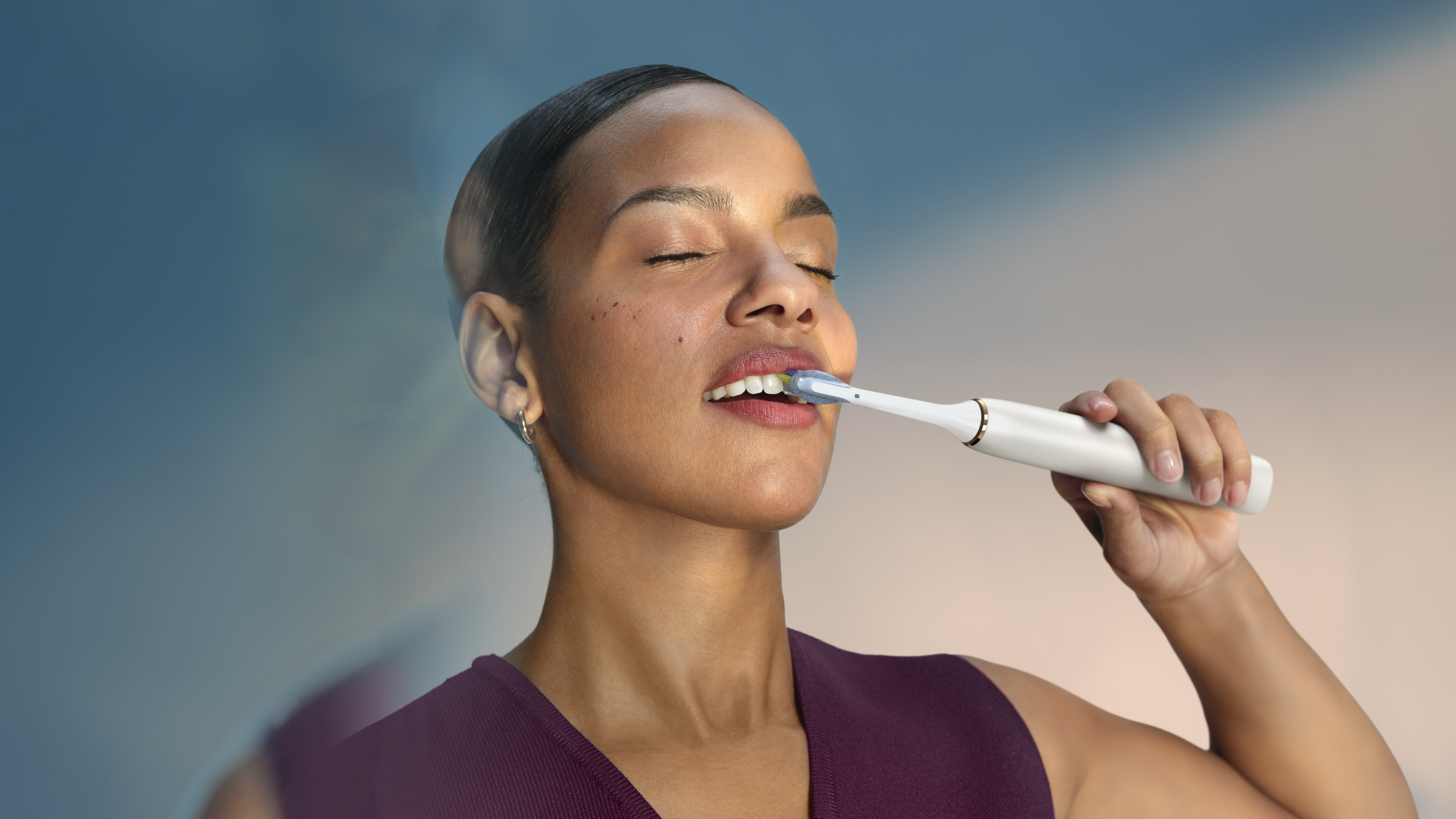 Una donna si lava i denti con uno spazzolino elettrico. Chiude gli occhi, con sfondo sfumato blu e arancione.