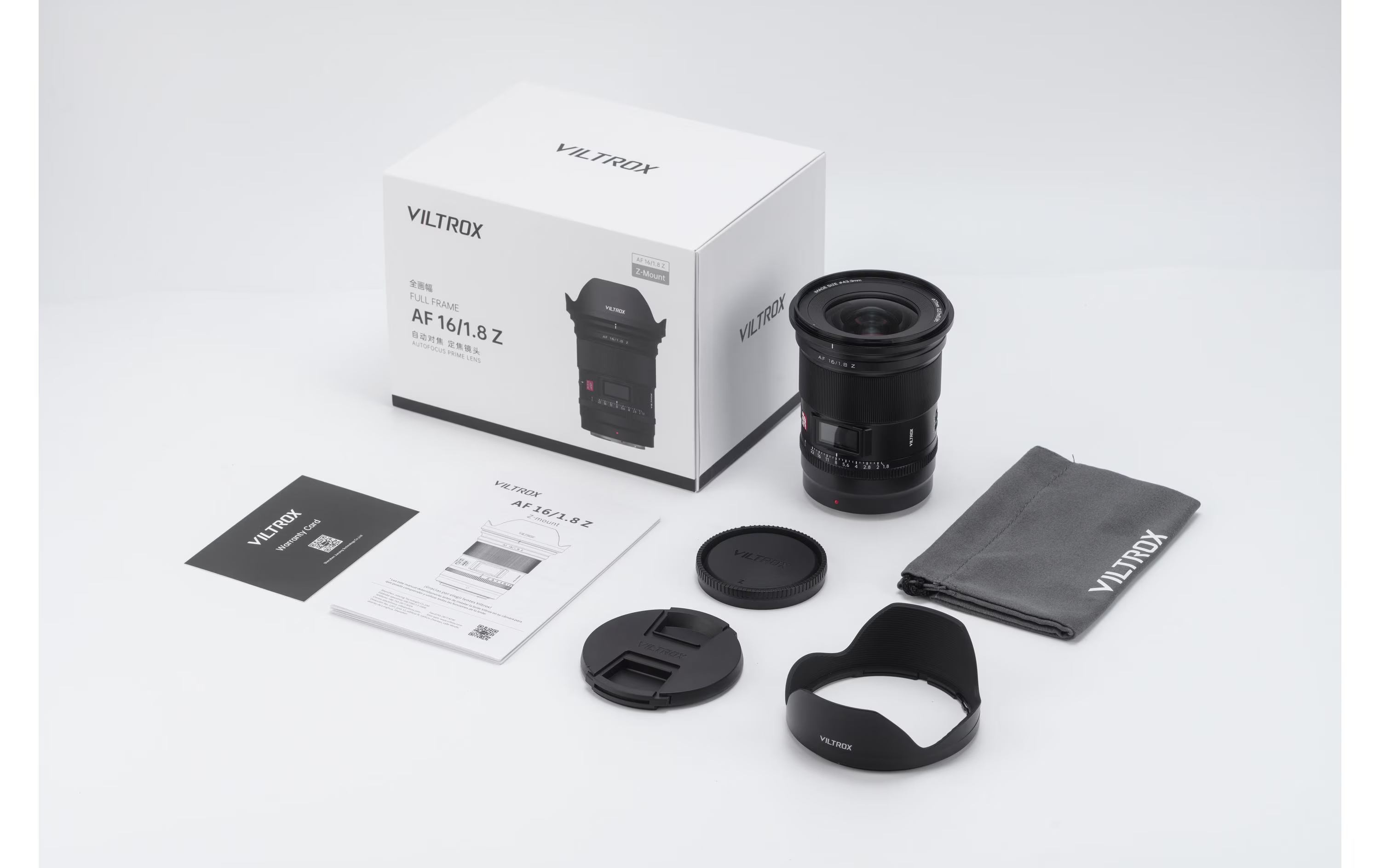 Ein Kameraobjektiv-Kit mit Schachtel, Objektiv, Gegenlichtblende, Kappen, Garantiekarte und Beutel, alles auf weißem Grund.