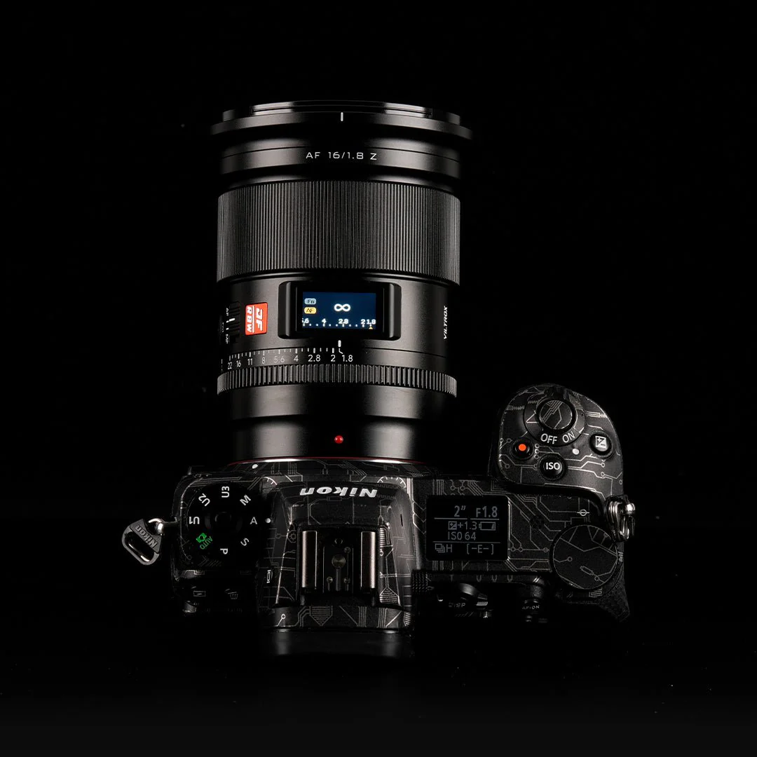 Eine schwarze Kamera mit Objektiv liegt auf einer schwarzen Oberfläche. Die Kamera zeigt Einstellungen an. Das Objektiv hat ein rot-weißes Etikett, Nikon.