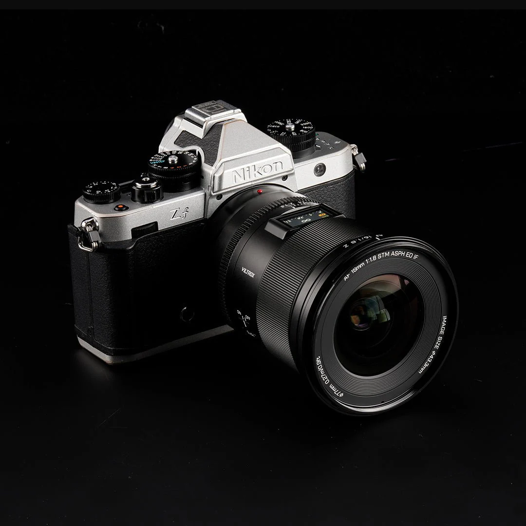 Eine Nikon Zf-Kamera mit schwarzem Objektiv liegt auf einer schwarzen Oberfläche. Die Kamera ist silber und schwarz, mit einem Objektiv von Viltrox.