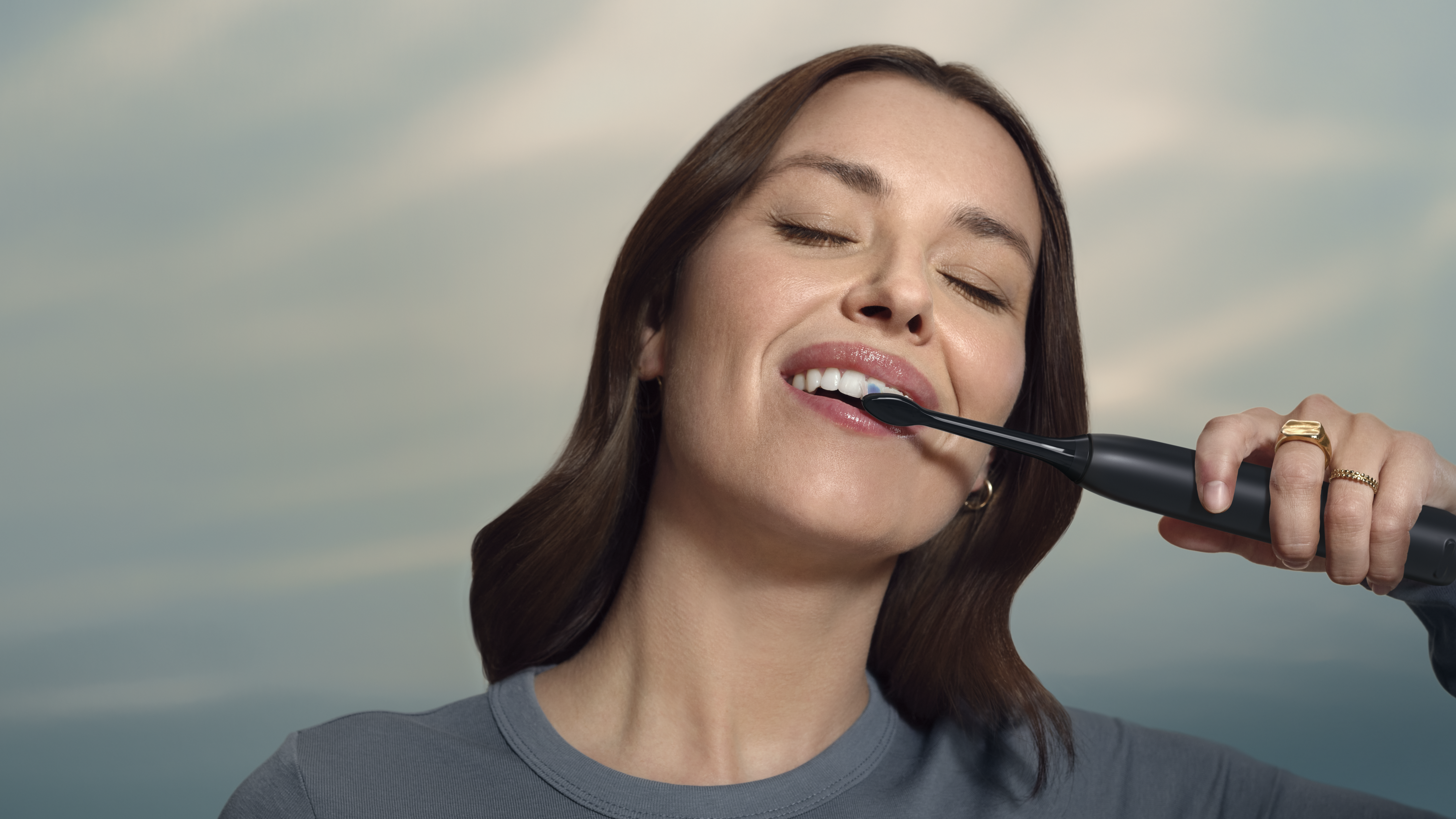 Donna che si lava i denti con uno spazzolino elettrico nero. Ha i capelli castani e indossa una maglietta grigia. Lo sfondo è sfocato.