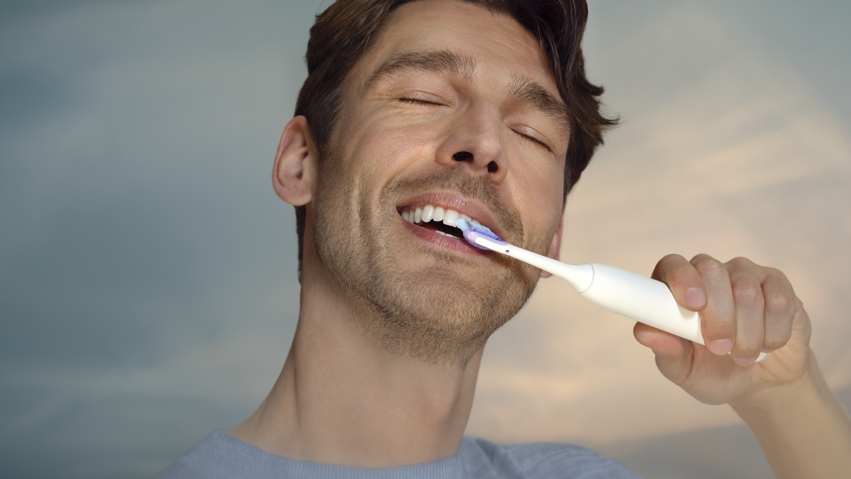 Un homme se brosse les dents les yeux fermés. Une brosse à dents électrique blanche est dans sa bouche. Le fond est un ciel flou.
