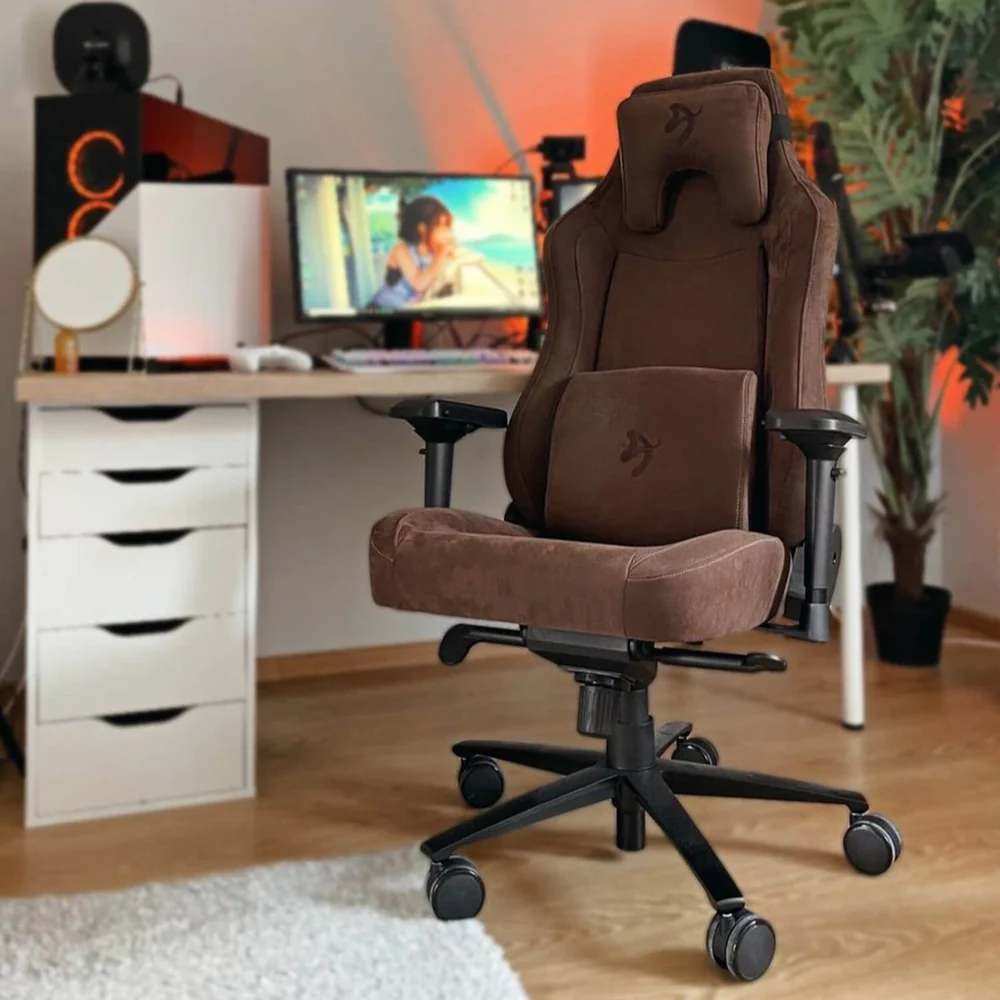 Egy barna gamer szék egy asztal előtt van, amin egy számítógép található. Balra egy fehér komód. A háttérben egy növény van.