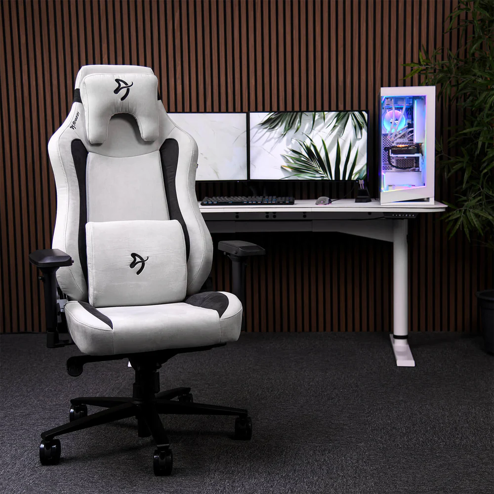 Egy gamer setup fehér-szürke székkel, két monitorral, számítógépházzal és asztallal. A háttérben fa burkolat látható.