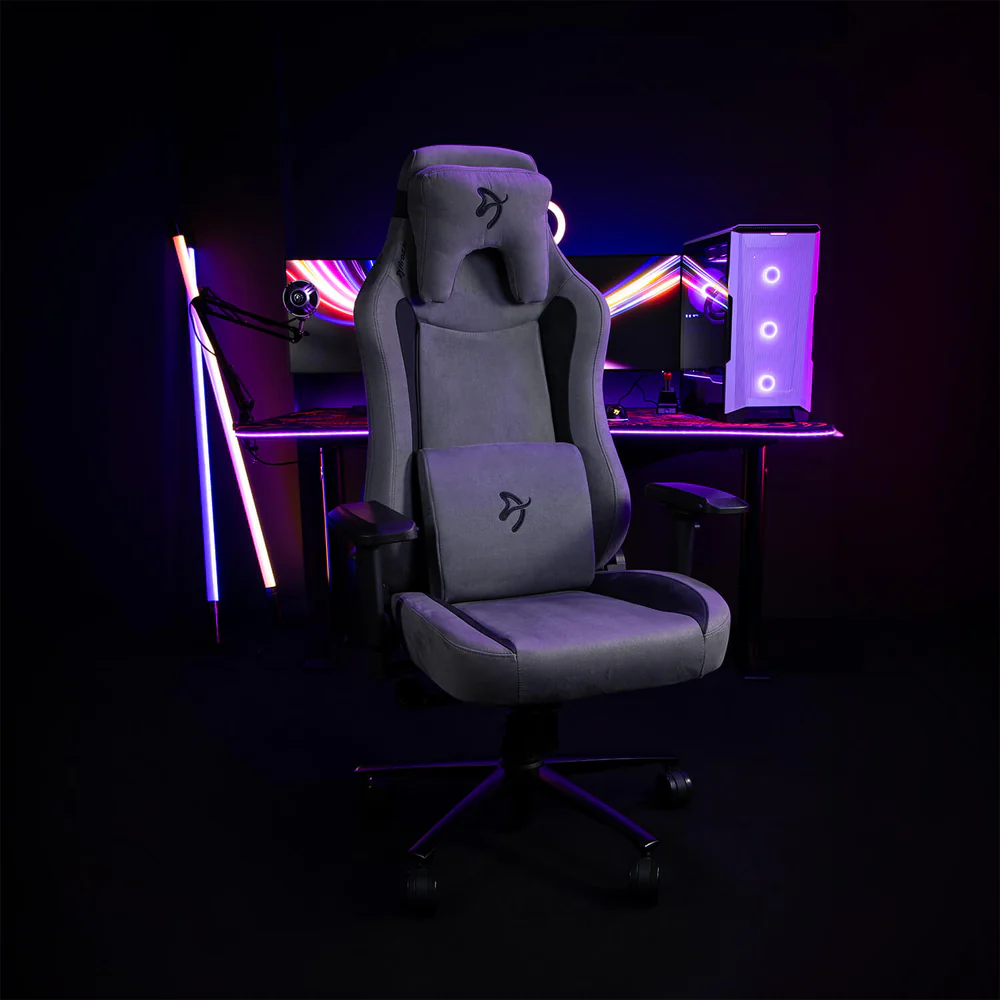 Egy szürke gamer szék egy asztal előtt, PC-vel és monitorral. Neonfények világítják meg a sötét szobát, hűvös hangulatot teremtve.