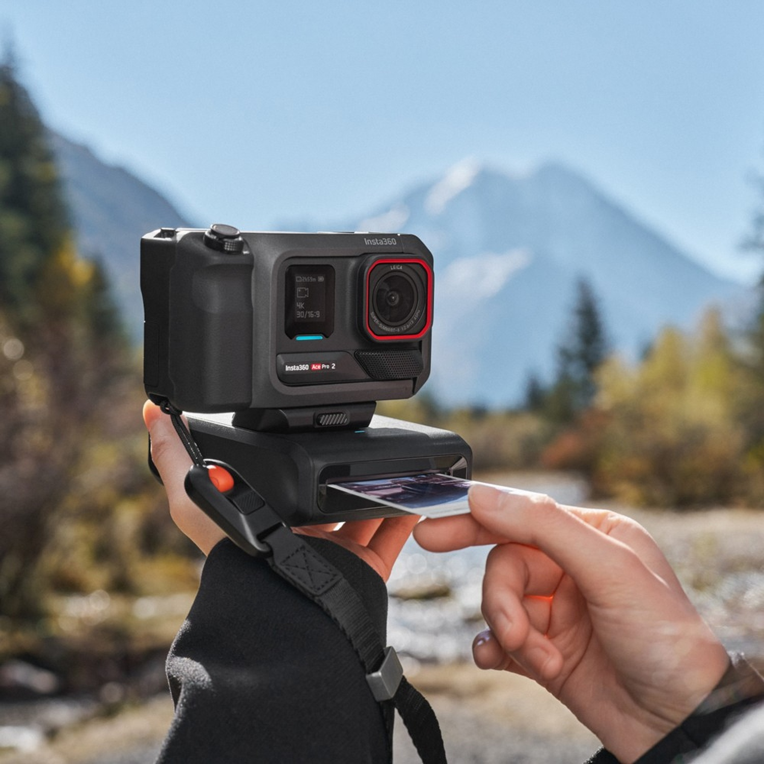 Una persona tiene in mano una fotocamera nera con lente rossa, stampando una foto. Montagne e alberi sullo sfondo.