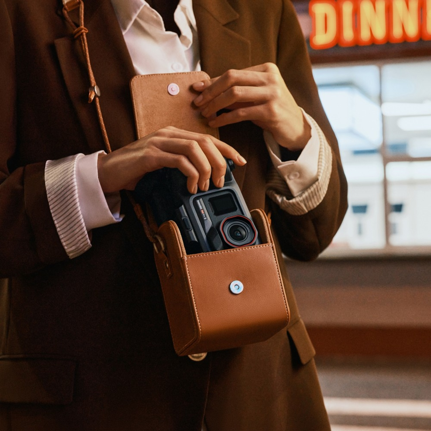 Una persona in completo marrone apre una borsa di cuoio marrone, rivelando una fotocamera nera con lente rossa. Sfondo con insegna 'DINNER'.