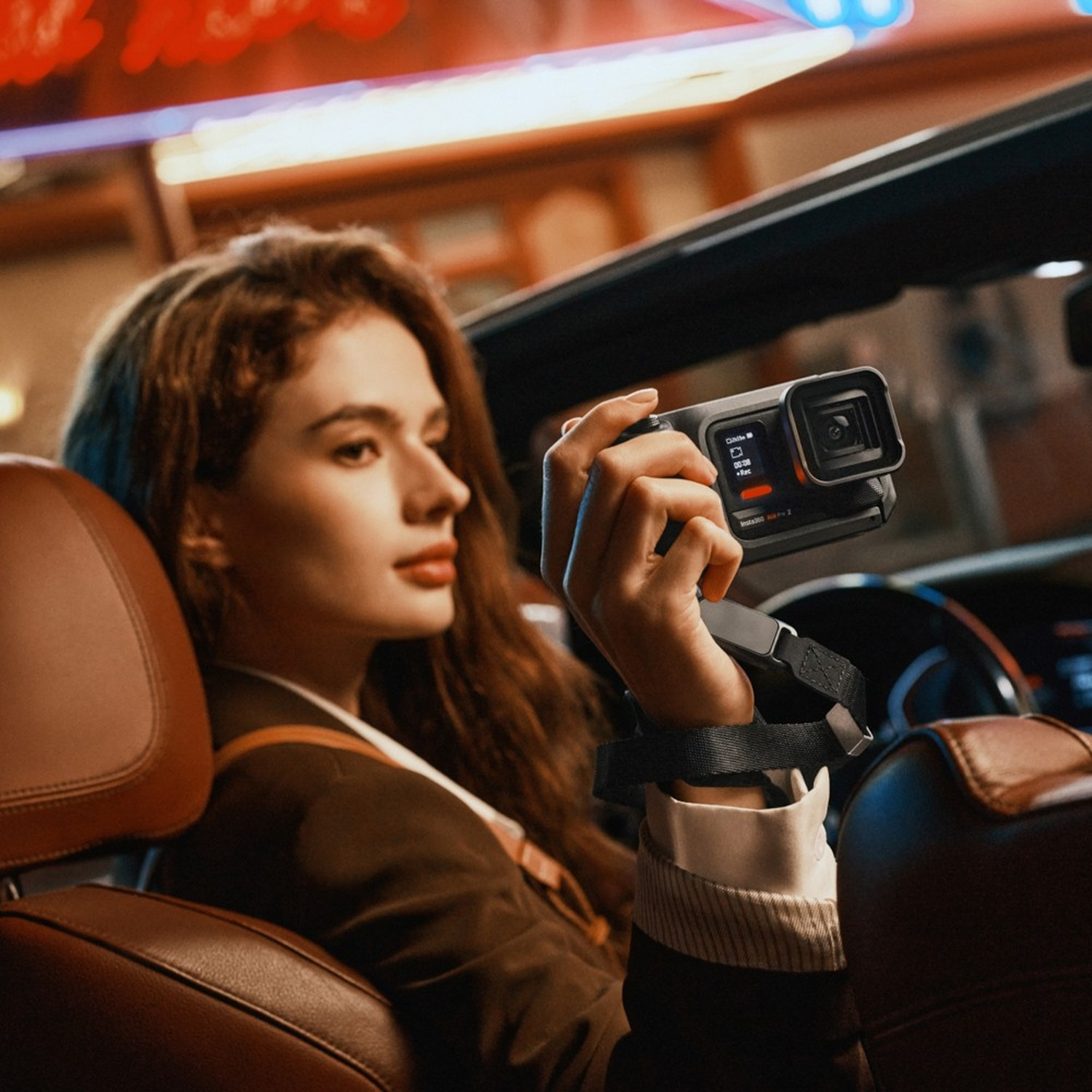 Una donna tiene una fotocamera in auto. Indossa una giacca marrone. Lo sfondo ha luci sfocate.