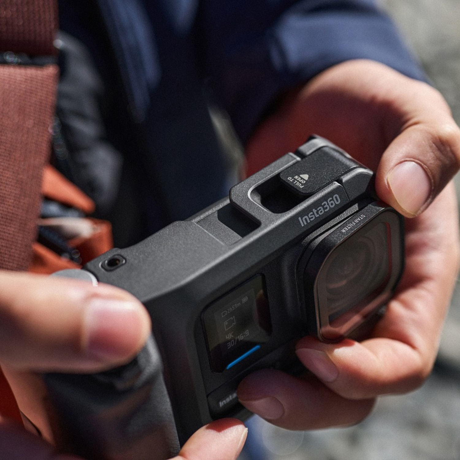 Una persona tiene in mano una fotocamera Insta360 nera. La fotocamera mostra le impostazioni su uno schermo. Lo sfondo è sfocato.