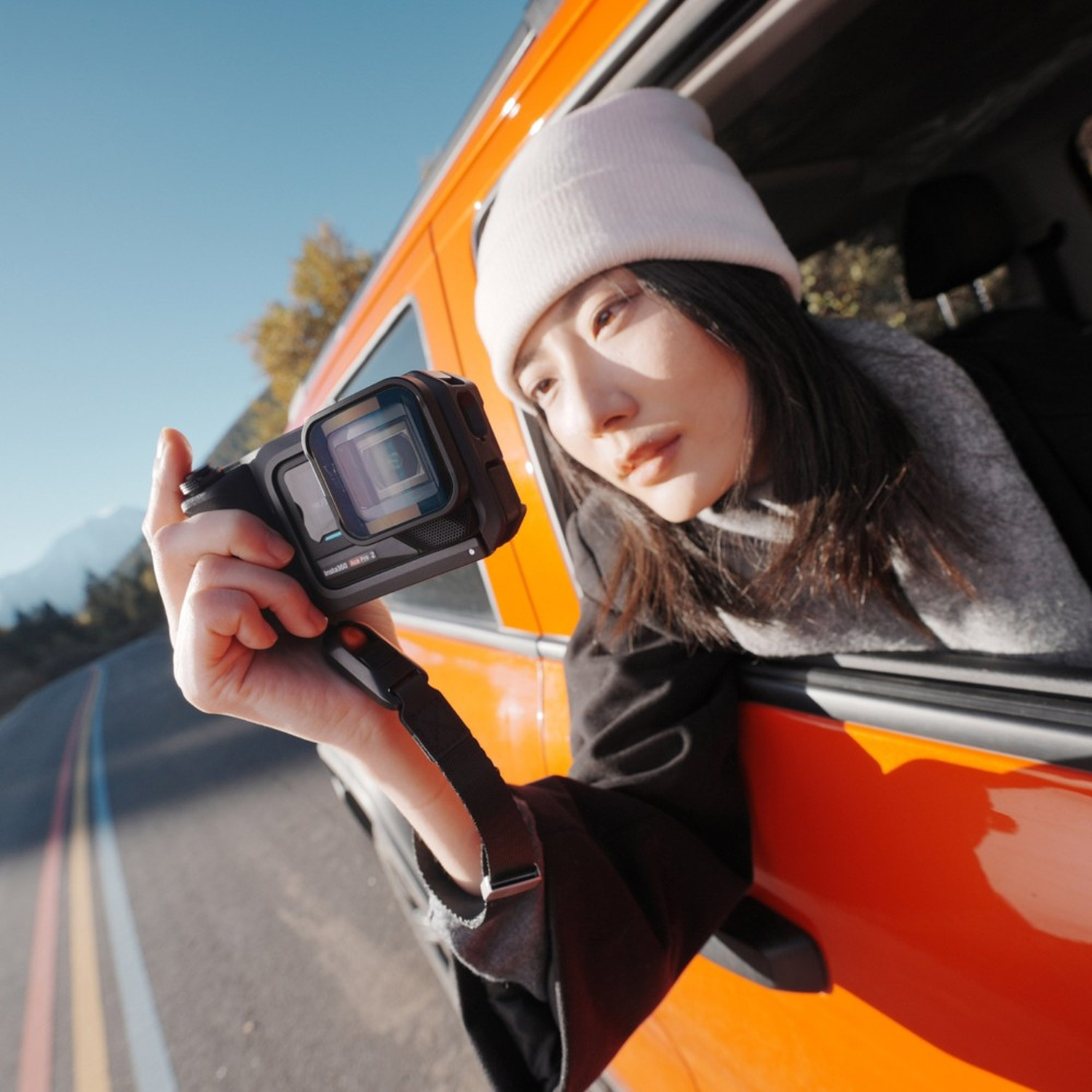 Una donna si sporge dal finestrino di un'auto arancione, tenendo in mano una fotocamera. Indossa un berretto e una giacca nera, catturando una scena stradale.