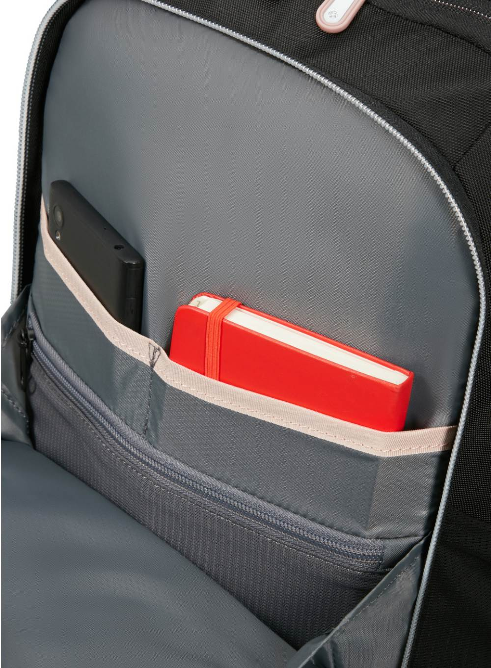 Wewnątrz czarnego plecaka: telefon i czerwony notatnik w przedniej kieszeni. Tło to szare wnętrze.