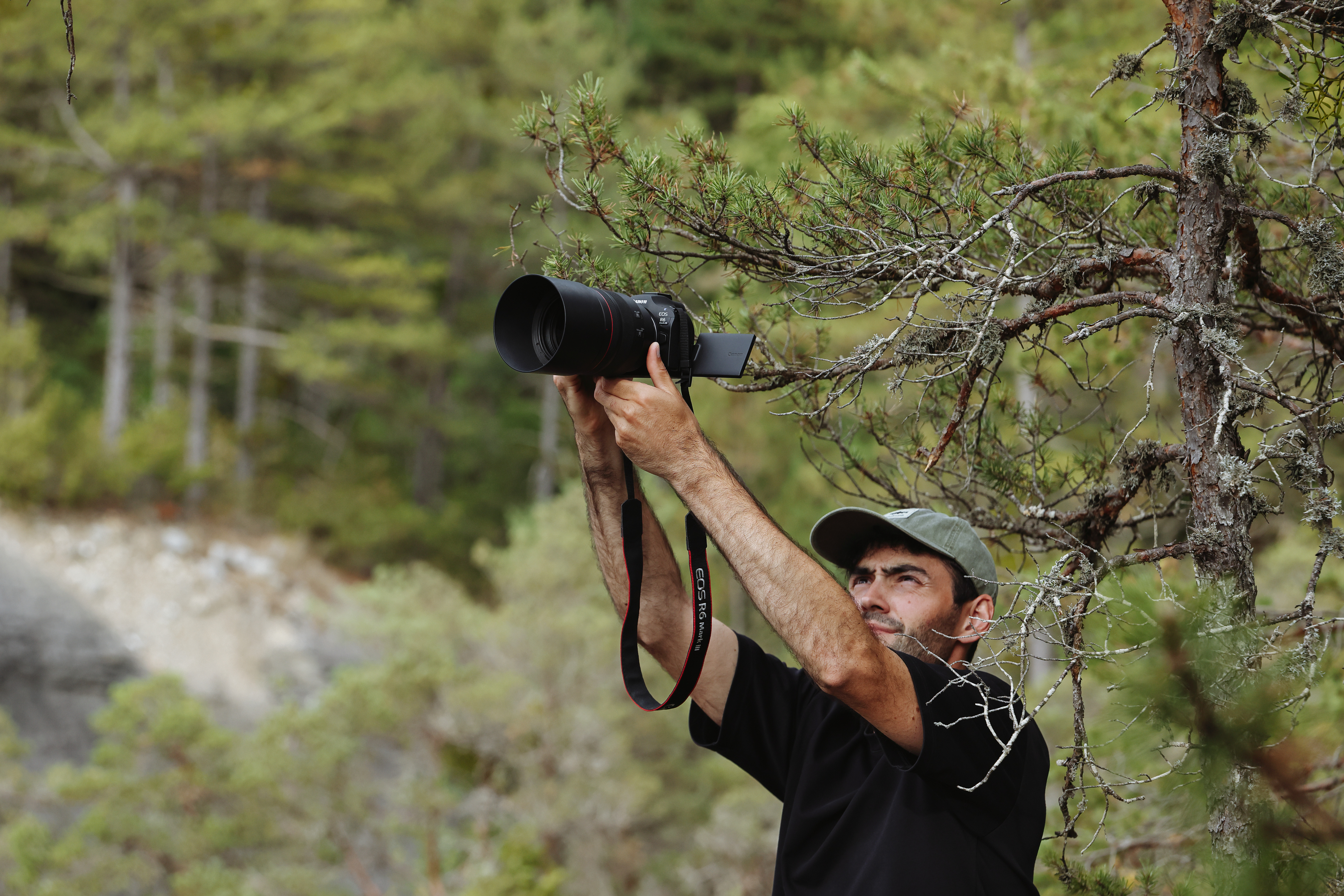 Ein Mann mit Kappe fotografiert die Natur mit einer Kamera mit großem Objektiv. Der Hintergrund zeigt Bäume und Laub.