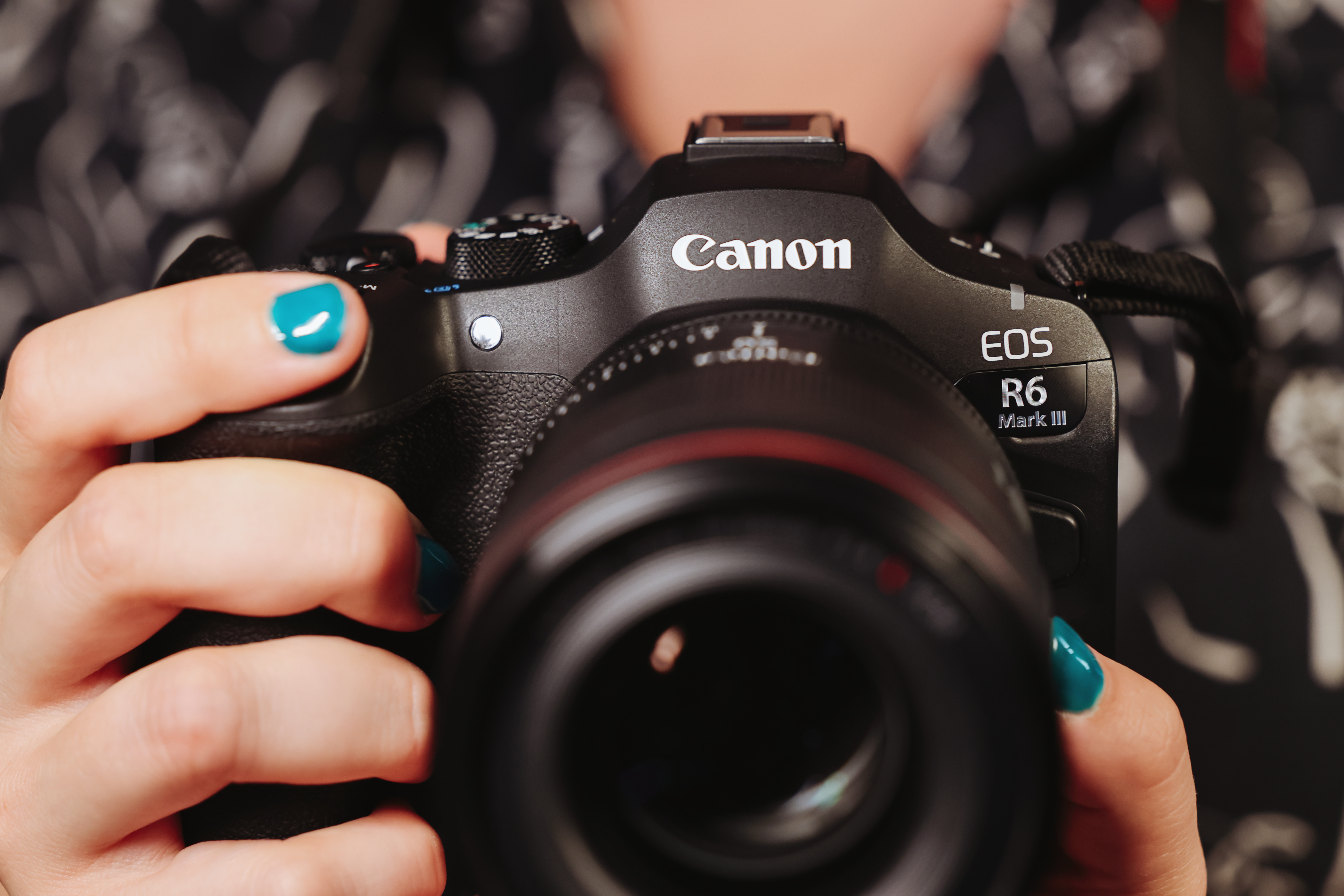 Eine Person hält eine Canon EOS R6 Mark III Kamera. Die Person hat türkisfarbenen Nagellack. Der Hintergrund ist unscharf.