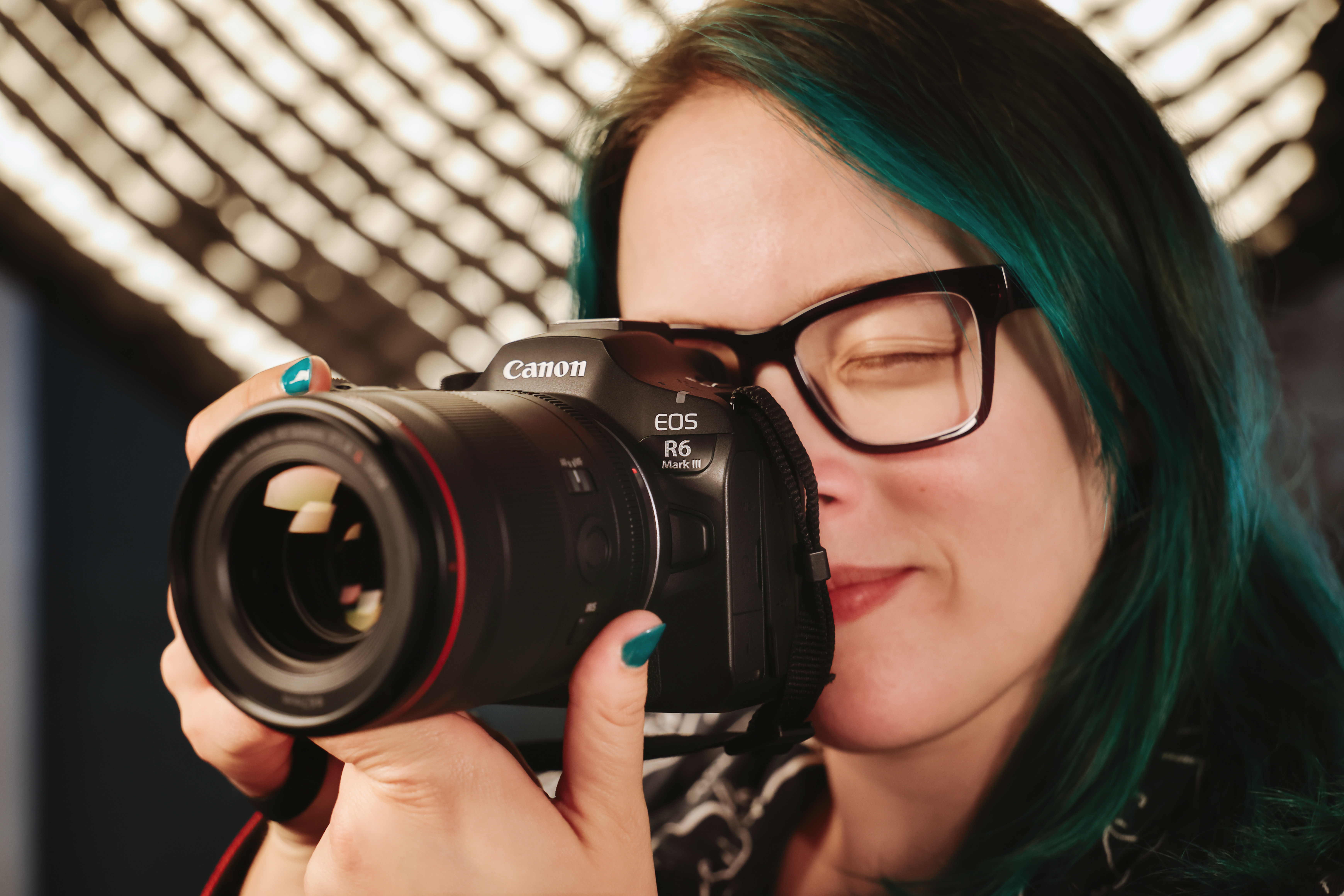 Frau mit grünen Haaren hält eine Canon-Kamera. Sie trägt eine Brille und hat türkisfarbenen Nagellack.