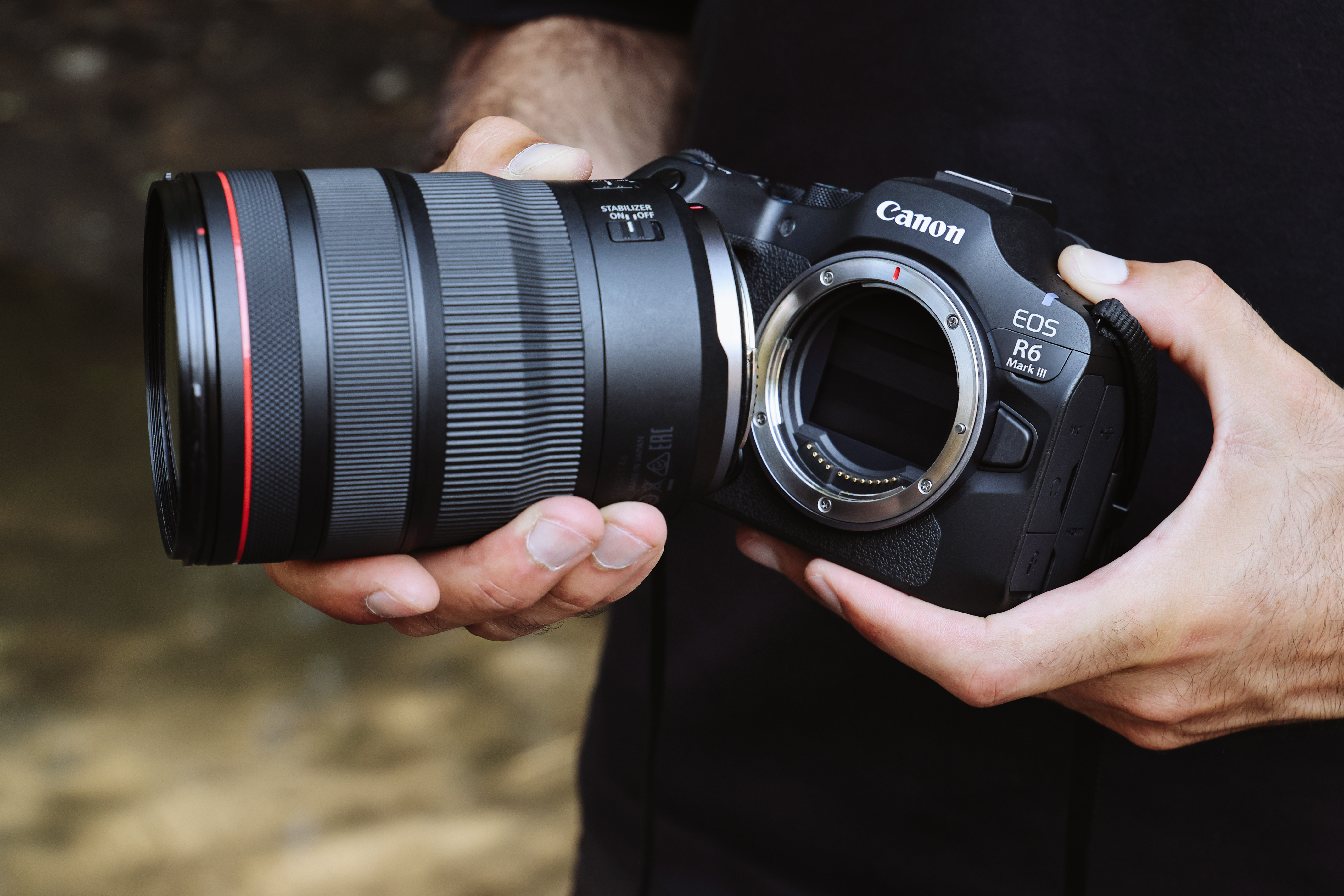Eine Person hält eine Canon EOS R6 Mark III Kamera mit einem Objektiv. Das Objektiv ist schwarz mit roten Akzenten.