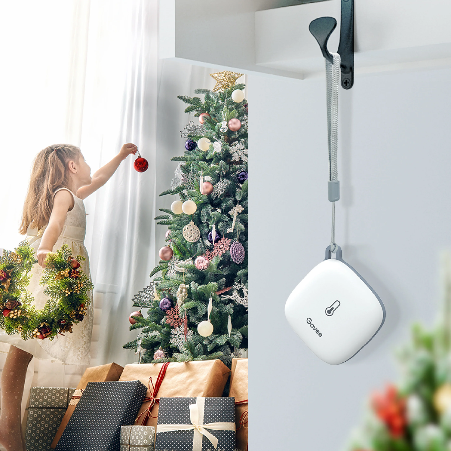 Ein Mädchen schmückt einen Weihnachtsbaum mit Ornamenten. Geschenke liegen unter dem Baum, ein Kranz in ihrer linken Hand. Ein weißes Gerät hängt.
