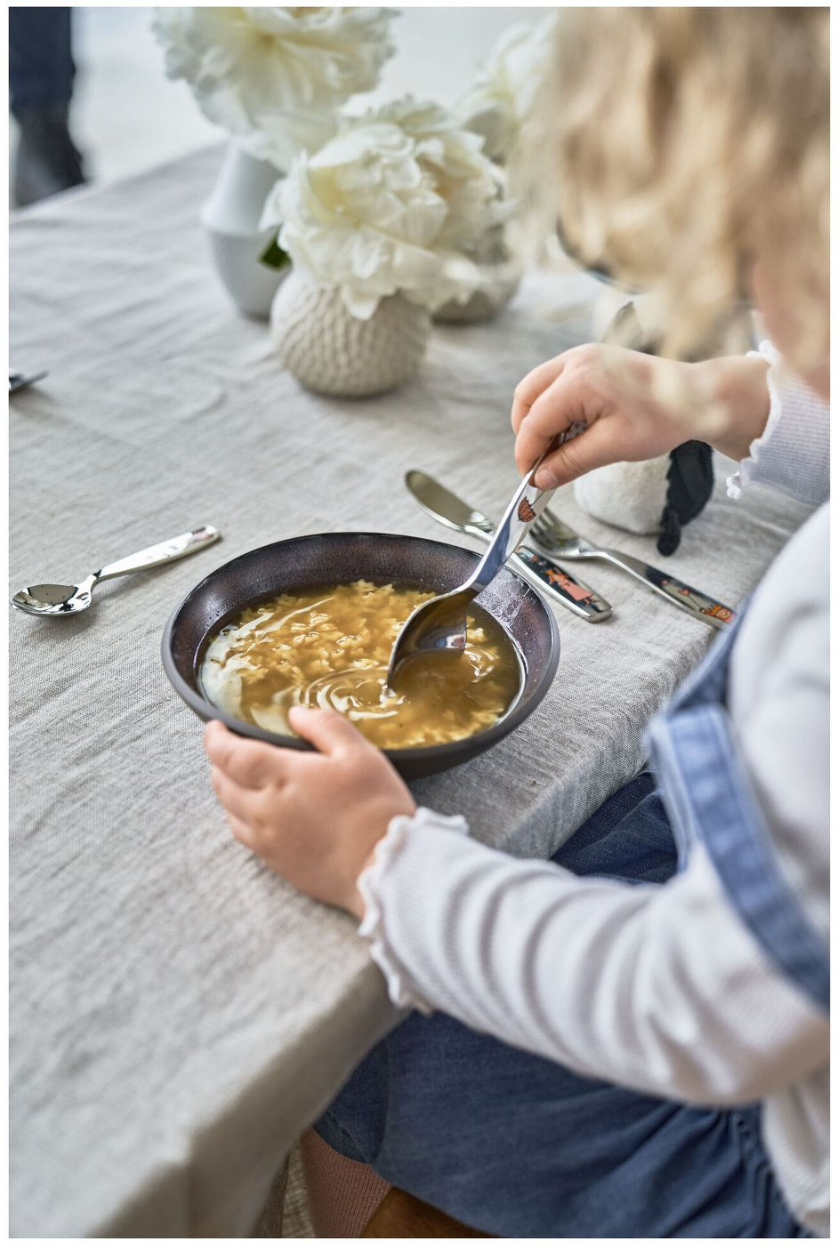 Ein Kind hält einen Löffel und schöpft Suppe aus einer Schüssel. Auf dem Tisch liegen weiße Blumen und Besteck. Das Kind trägt ein weißes Hemd und blaue Latzhosen.