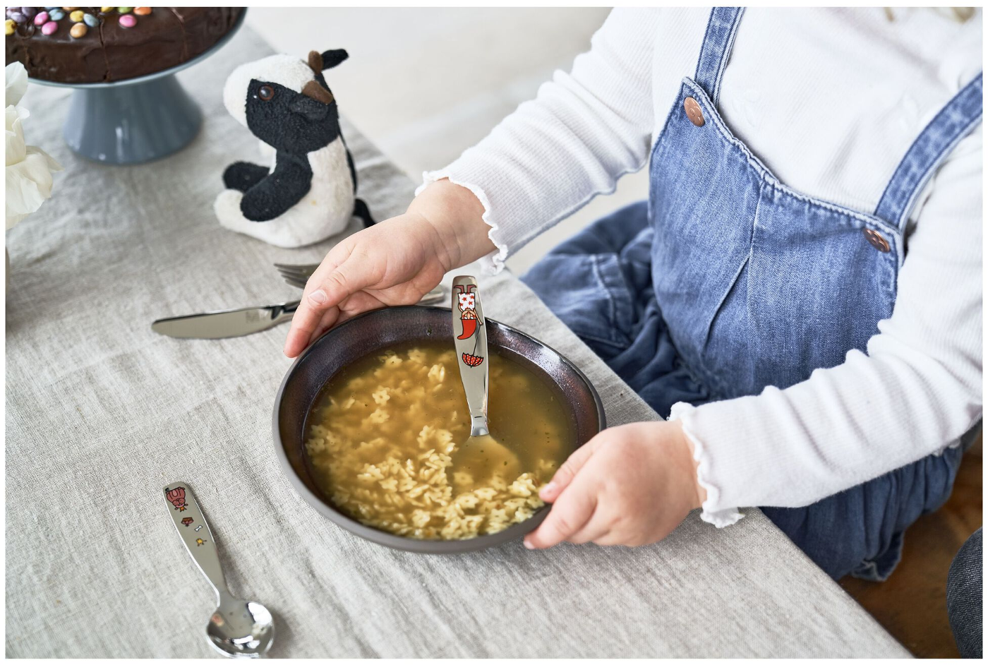 Ein Kind hält eine Schüssel mit Suppe mit sternförmiger Pasta, einem Löffel und einem Plüschkuh auf einem Tisch.