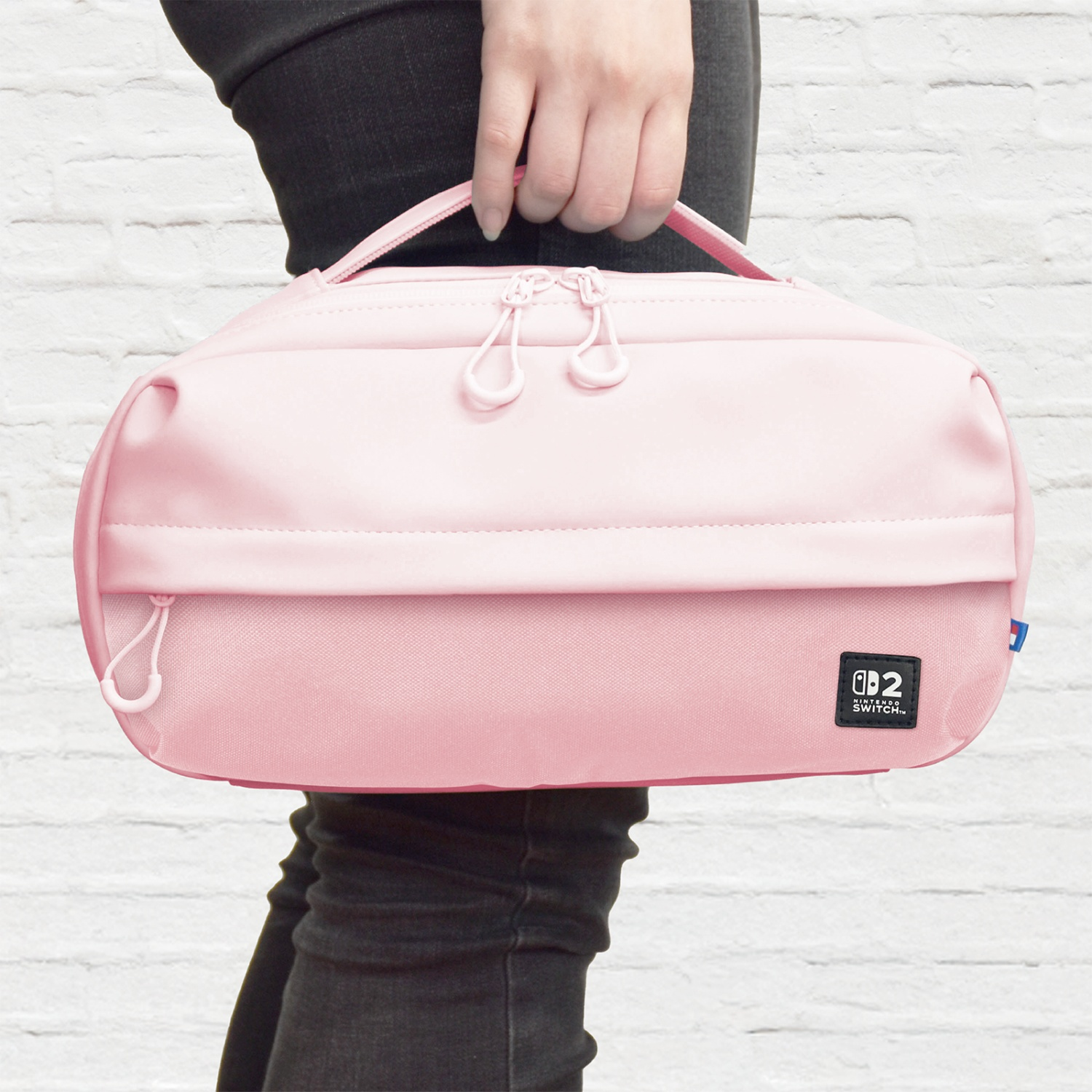Una persona tiene in mano una borsa Nintendo Switch rosa. La borsa ha una cerniera, un manico e un logo Nintendo Switch. Lo sfondo è un muro di mattoni bianchi.