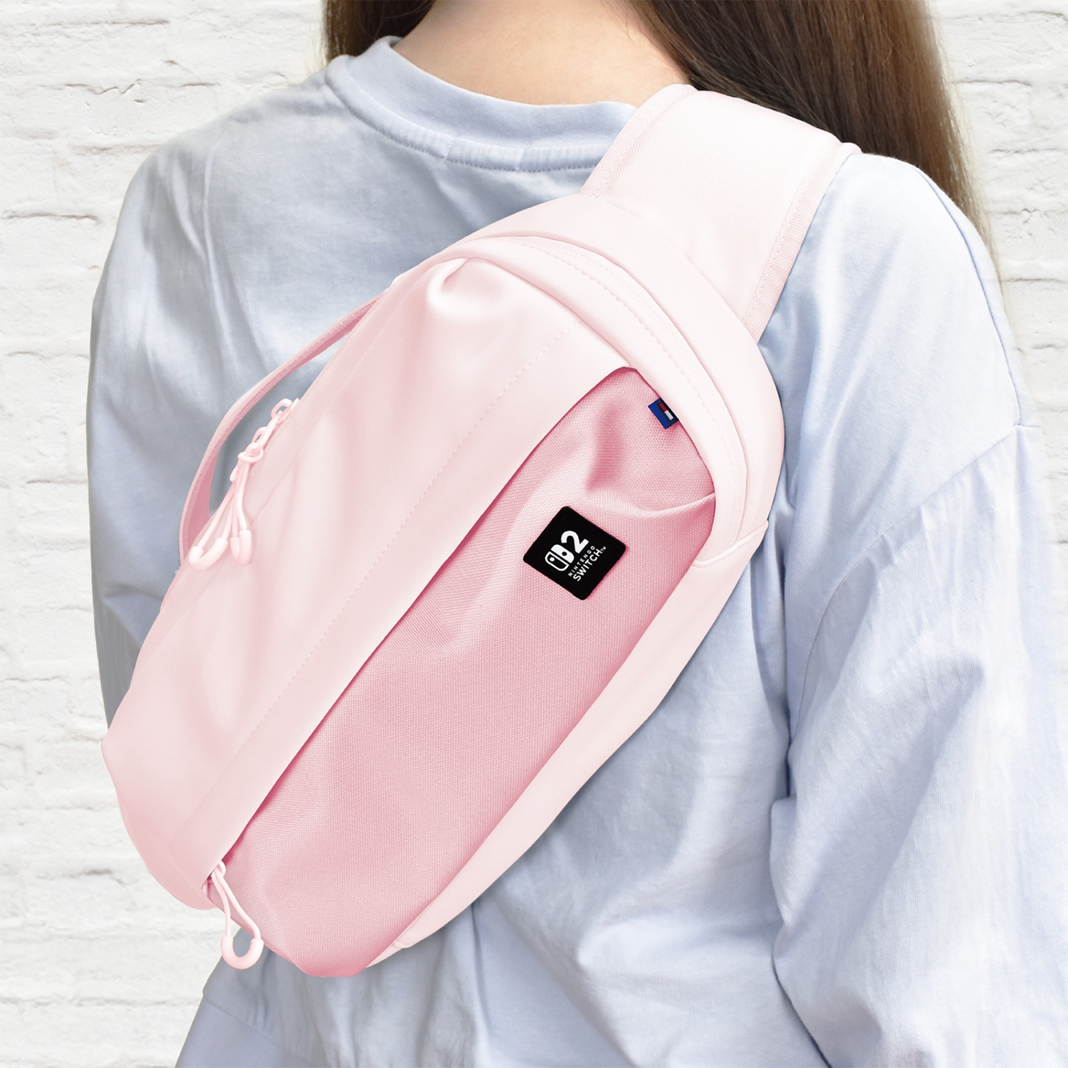 Una donna indossa una borsa a tracolla rosa chiaro su una camicia azzurra. La borsa ha un logo Nintendo Switch. Lo sfondo è un muro di mattoni bianchi.
