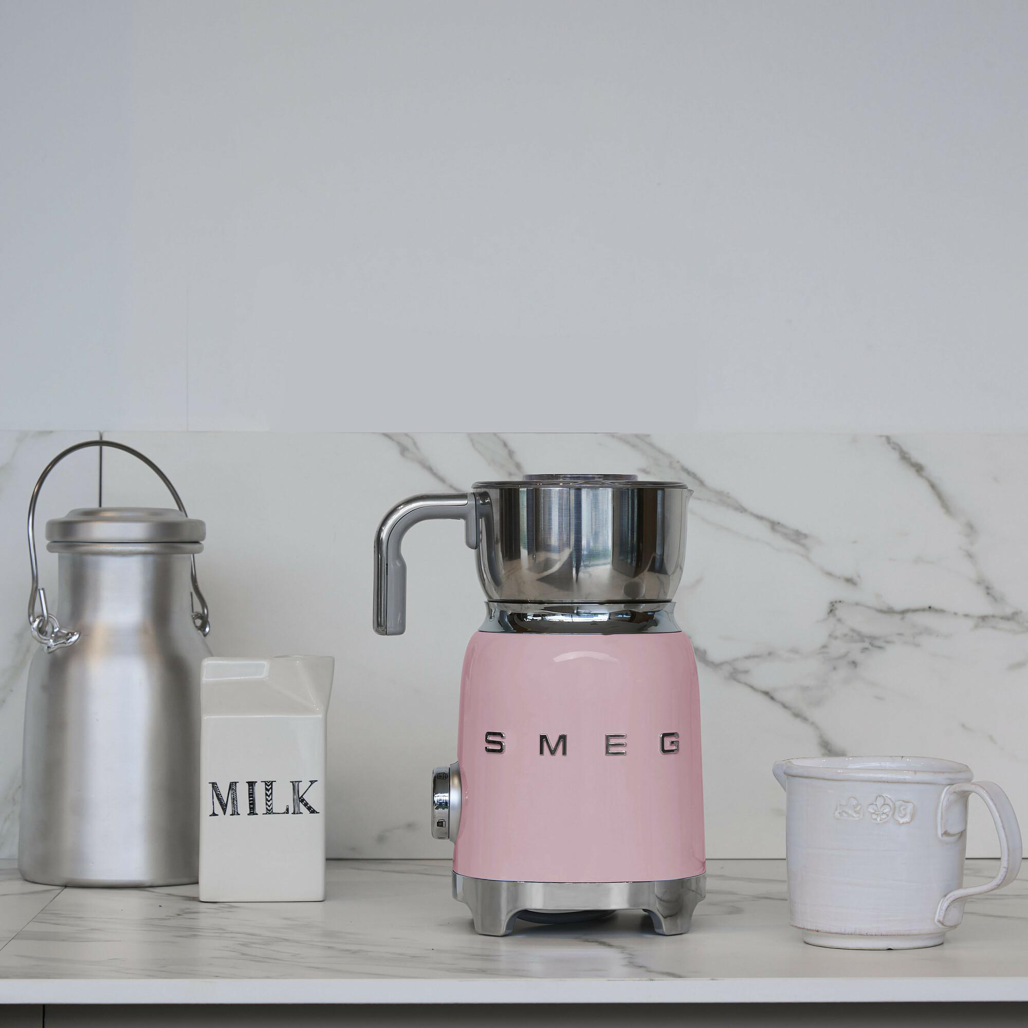 Rosa SMEG Milchaufschäumer, Milchkanne aus Metall, Milchkarton und eine weiße Tasse auf Marmorplatte.