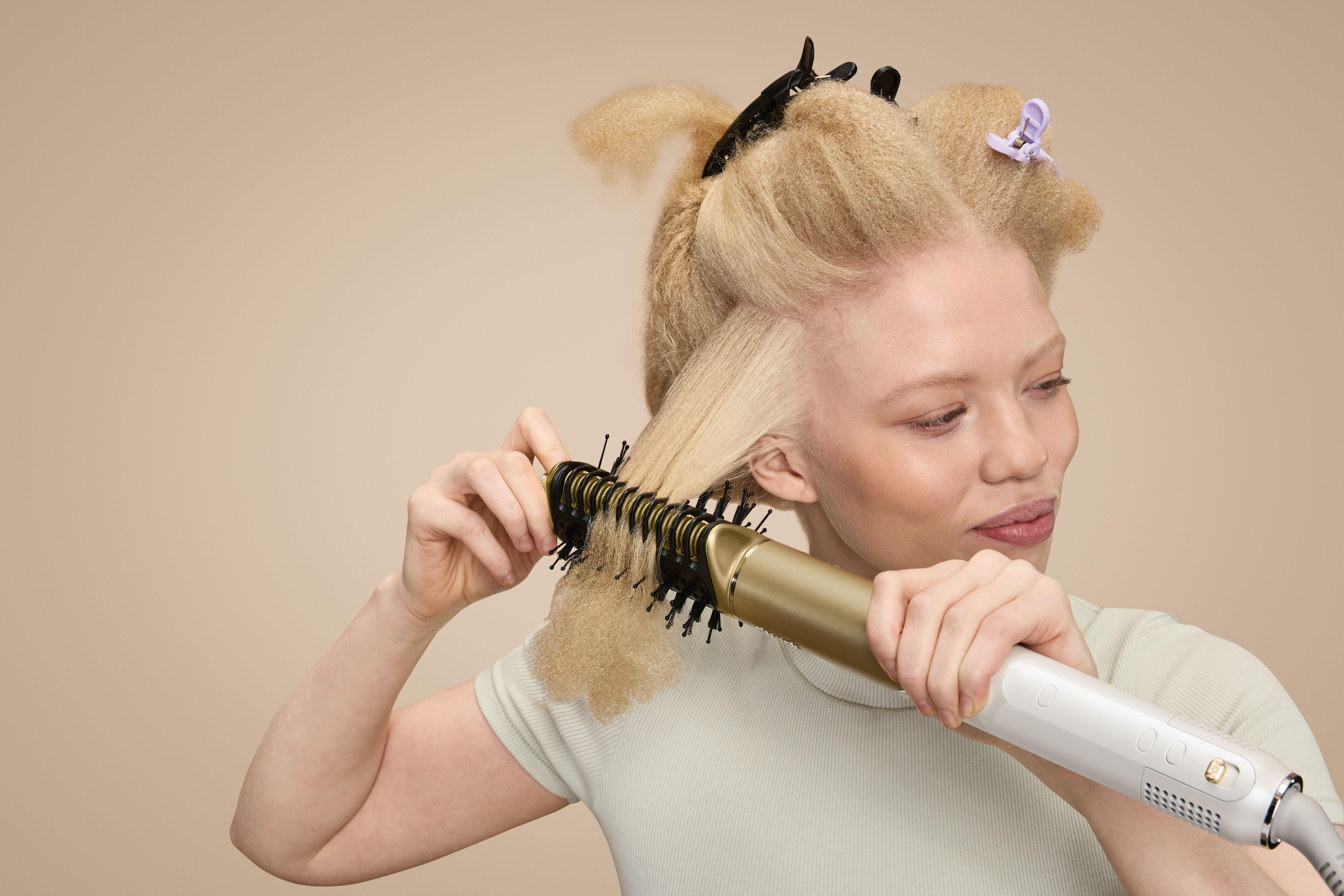 Eine Frau mit blondem Haar stylt ihr Haar mit einem gold-schwarzen Haarstyling-Tool. Haarklammern sind in ihrem Haar.