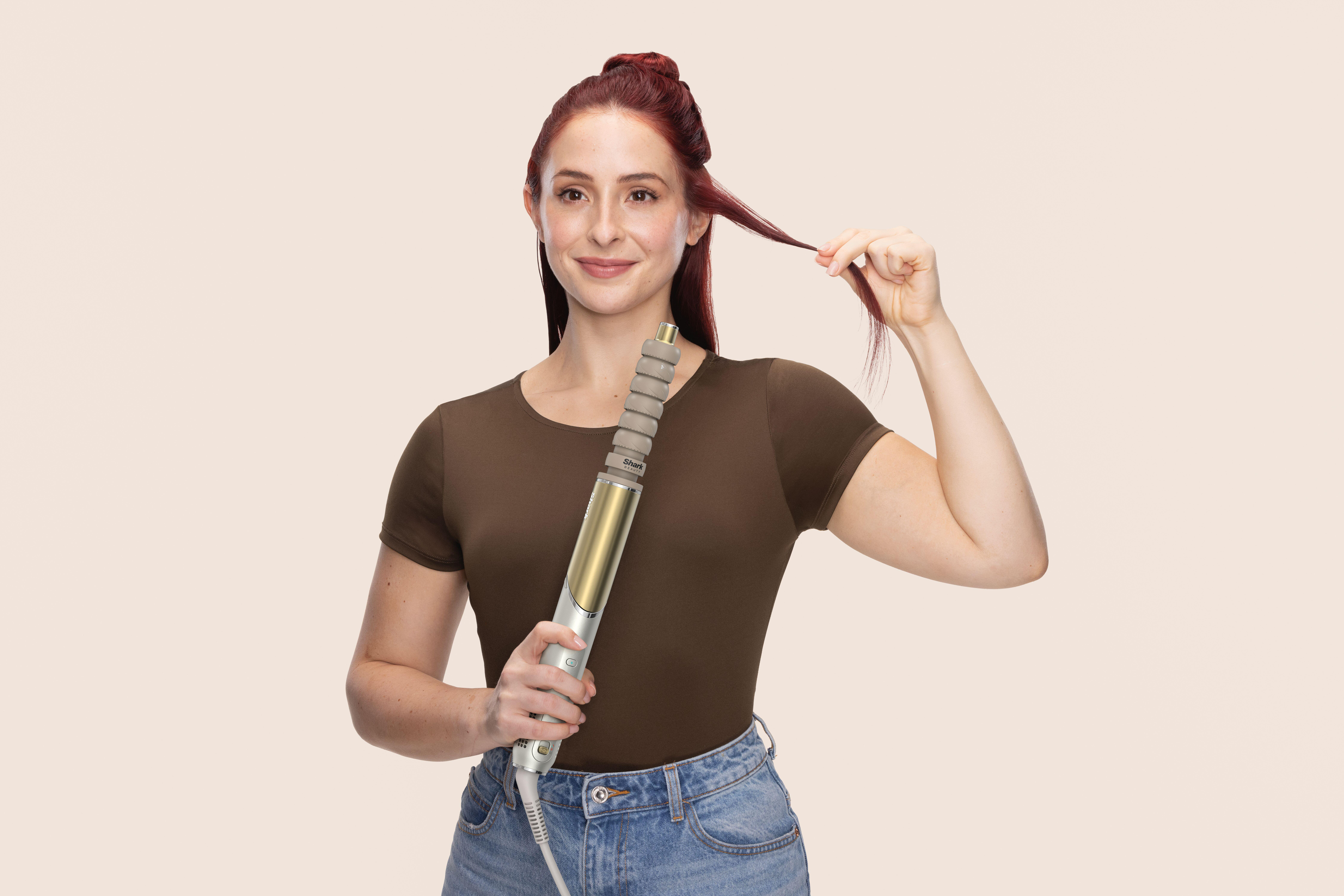 Frau hält ein Haarstyling-Tool, mit roten Haaren und braunem Hemd, vor beigem Hintergrund.