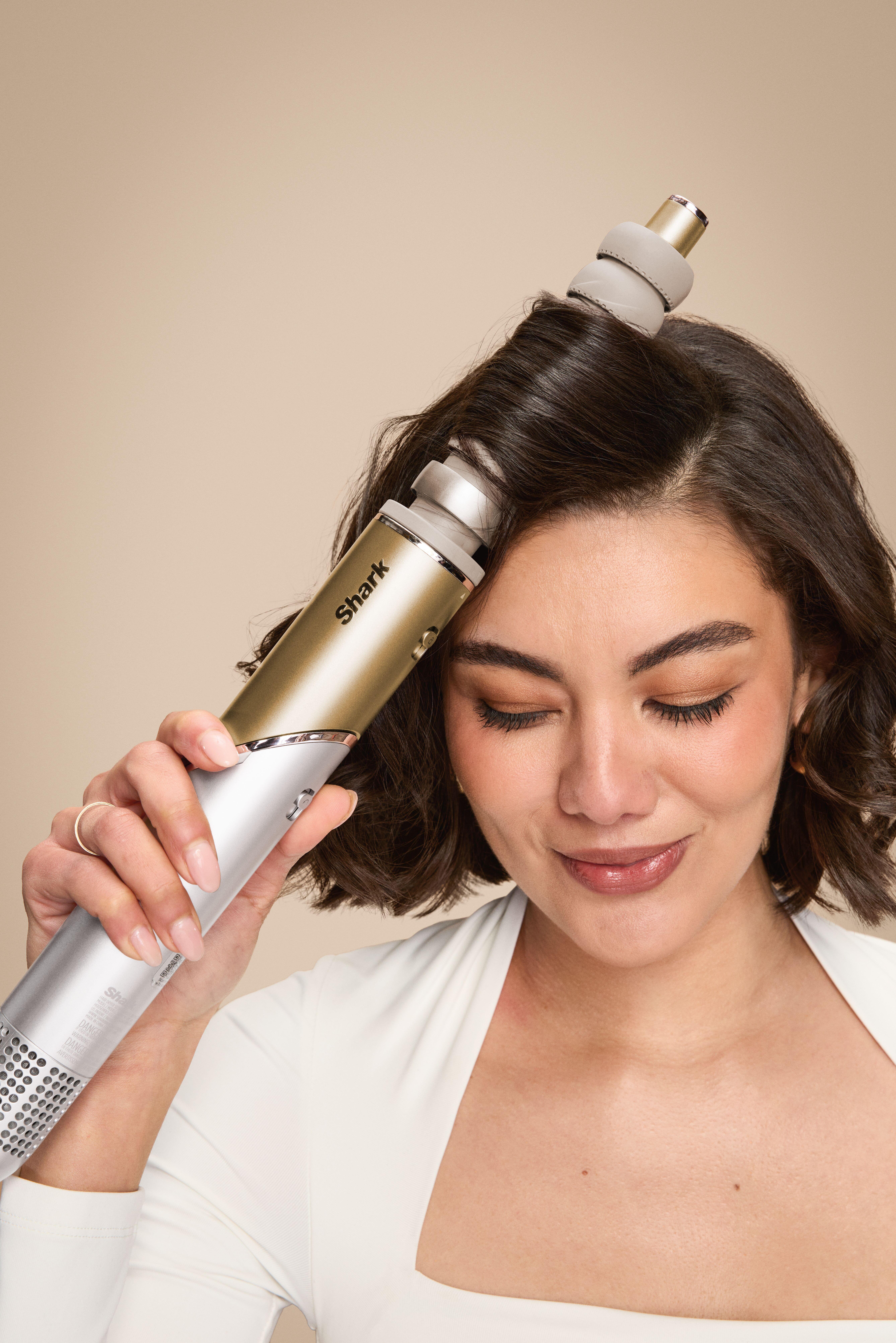 Eine Frau benutzt einen Shark-Haarstyler. Sie hält ihn an ihr Haar, die Augen geschlossen, mit einem Gold- und Silberdesign. Der Hintergrund ist beige.