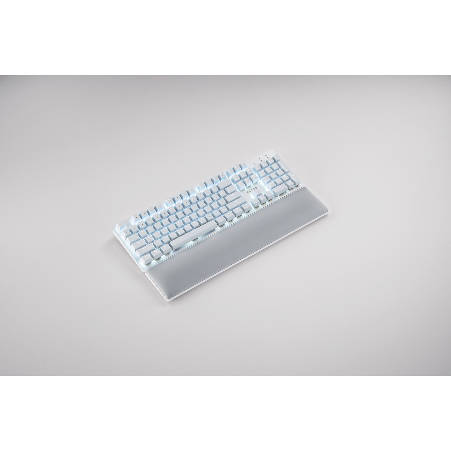 A white Razer keyboard with illuminated keys and a wrist rest lies on a gray surface. The background is plain gray.