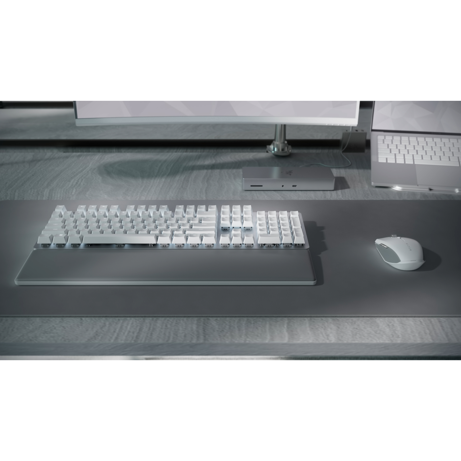 A white keyboard, mouse, and other computer peripherals on a desk. A monitor and laptop are in the background.